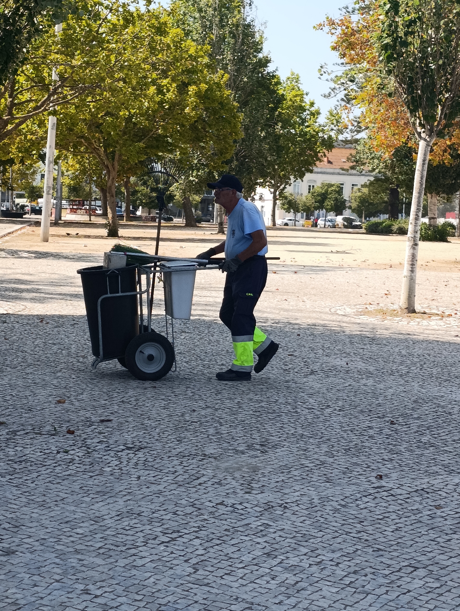 A man wearing a blue shirt and dark pants with fluorescent yellow leg bands is pushing a cleaning cart on a cobblestone path in Setúbal, Portugal. The cart has two black bins, one white bin, and a broom attached. He is walking from right to left in the frame. In the background, there are trees with green and yellow leaves, and a building with a red roof. The sky is clear and blue.
