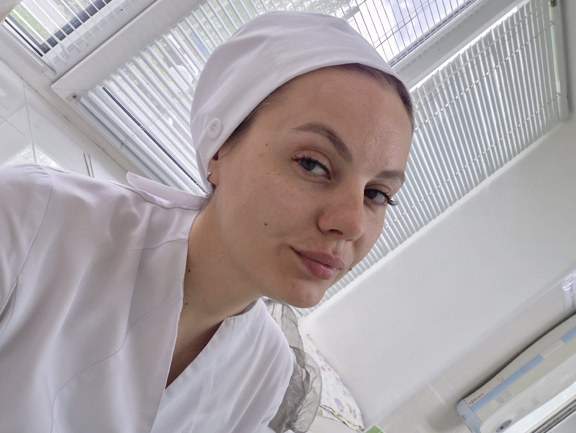 A woman in a white medical cap and white coat is looking down at the camera. She is in what appears to be a medical or clinical setting. The background includes a white tiled wall, a window with blinds, and what appears to be medical equipment or supplies. It's not possible to confirm if the image was taken in Chaikovsky, Russia, based on the visual information provided.