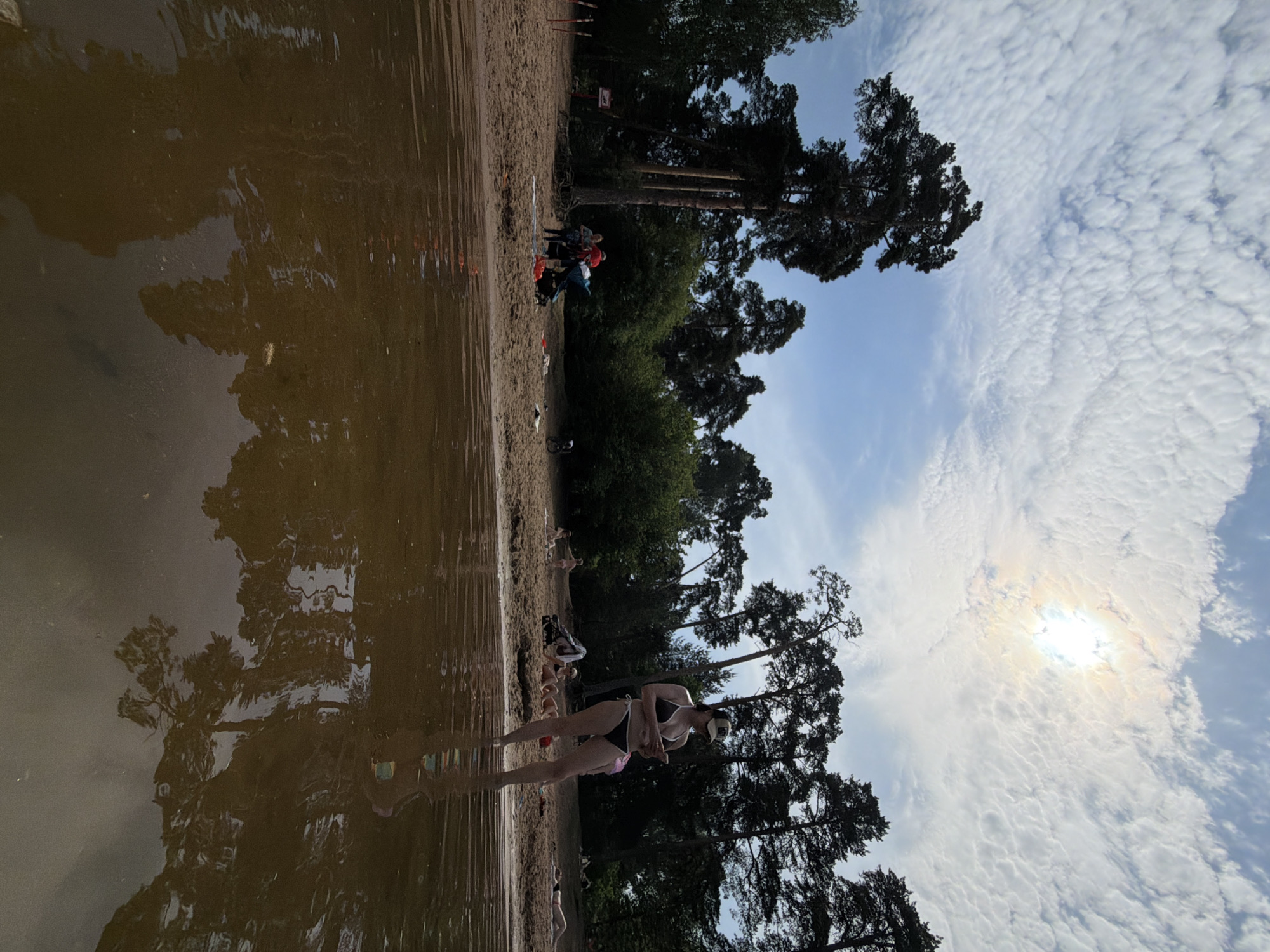 The image shows a beach scene, likely in Moscow, Russia, based on the presence of a typical sandy shoreline. The foreground features shallow water reflecting the trees and sky, creating a mirrored effect. In the water, a woman is standing in a bikini and cap. Other individuals are visible on the sandy beach, some sitting or standing. Behind them, there are trees and a blue sky with clouds. The scene gives the impression of a summer day at a recreational area.