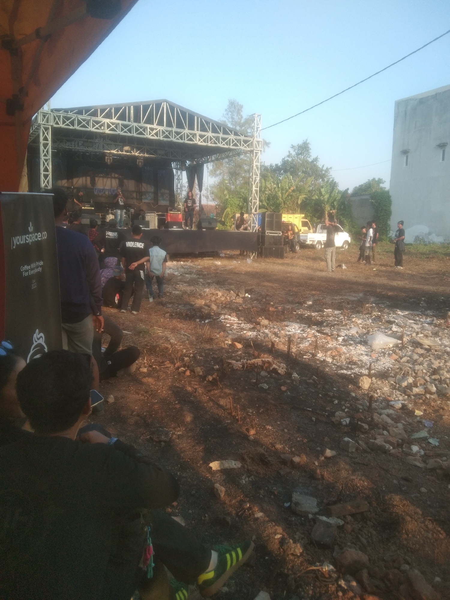 The image depicts an outdoor event with a stage set up for a performance, likely a concert, in a rural or undeveloped area. In the foreground, several people are visible, some seated and others standing, looking towards the stage. The ground is uneven and covered with dirt, rocks, and debris, suggesting an unpaved lot or construction site.
The stage is constructed from metal scaffolding and covered with a large backdrop. Several musicians are on stage, one of whom appears to be singing into a microphone. A sound system with large speakers is set up in front of the stage.
In the background, a white building with two windows is visible, along with some trees and vegetation. A yellow truck is parked behind the stage area. The sky is clear and blue, indicating daytime. The overall scene suggests a casual, outdoor gathering focused on live music.
While the image doesn't explicitly state the location, the provided context states it is in Kecamatan Haurgeulis, Indonesia. This implies the scene is taking place in that specific sub-district.