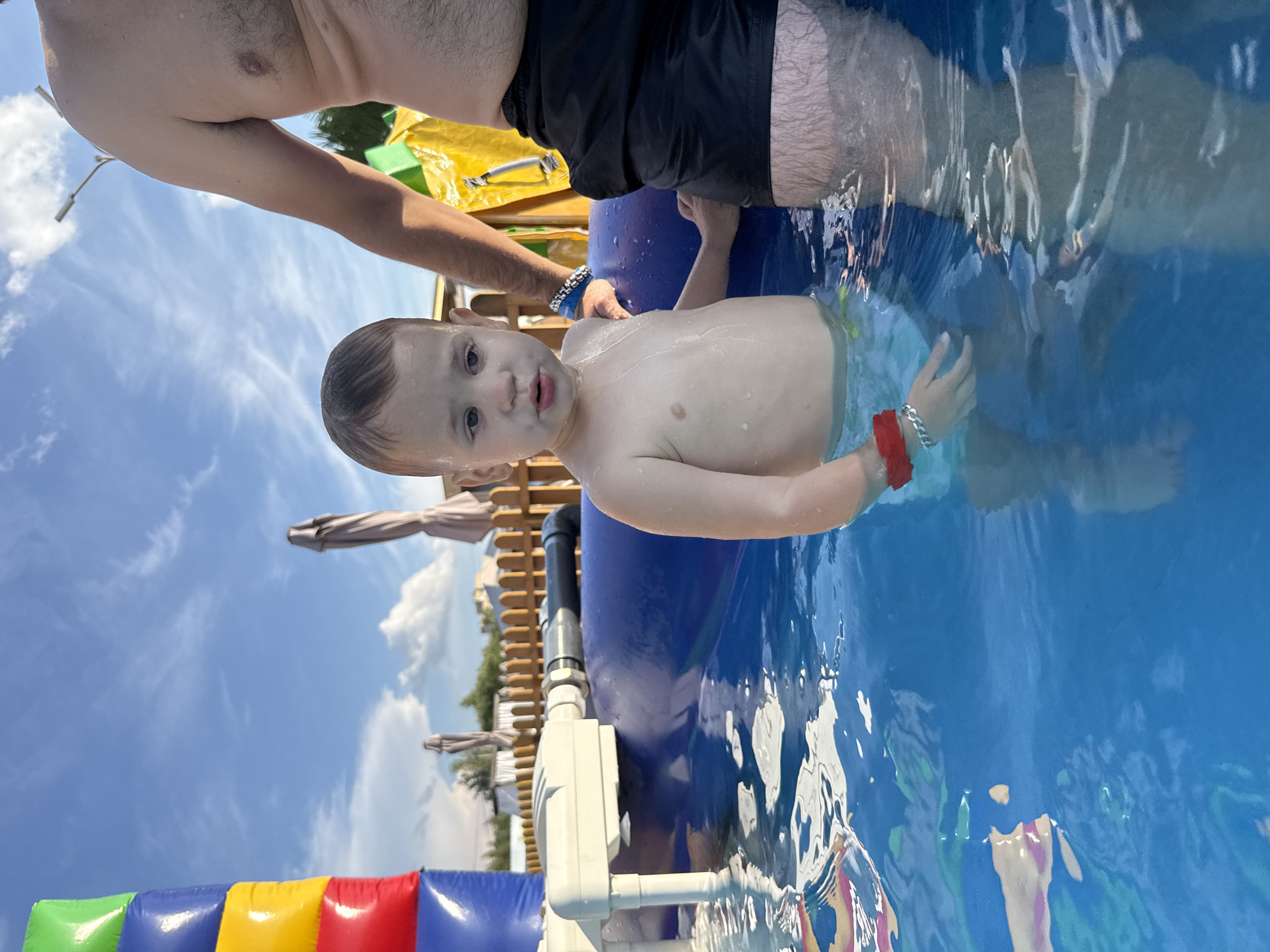 An outdoor, medium shot captures a young Caucasian child standing in a shallow, blue inflatable pool on a sunny day. The child, with light skin and brown hair, is wearing a red wristband and a light-colored diaper or swim trunks. The child is looking towards the viewer with a slightly open mouth, and water droplets are visible on their skin. To the right of the child, an adult's torso is visible, wearing black swim shorts. The adult's arm is extended, and they are wearing a blue bracelet. In the background, a tall, inflatable structure with colored blocks of blue, yellow, red, and green stands on the left. Several closed beach umbrellas and a wooden fence are also visible, along with a light pole with a fixture. The sky above is blue with scattered white clouds. The water in the pool has reflections of the sky and surrounding objects.