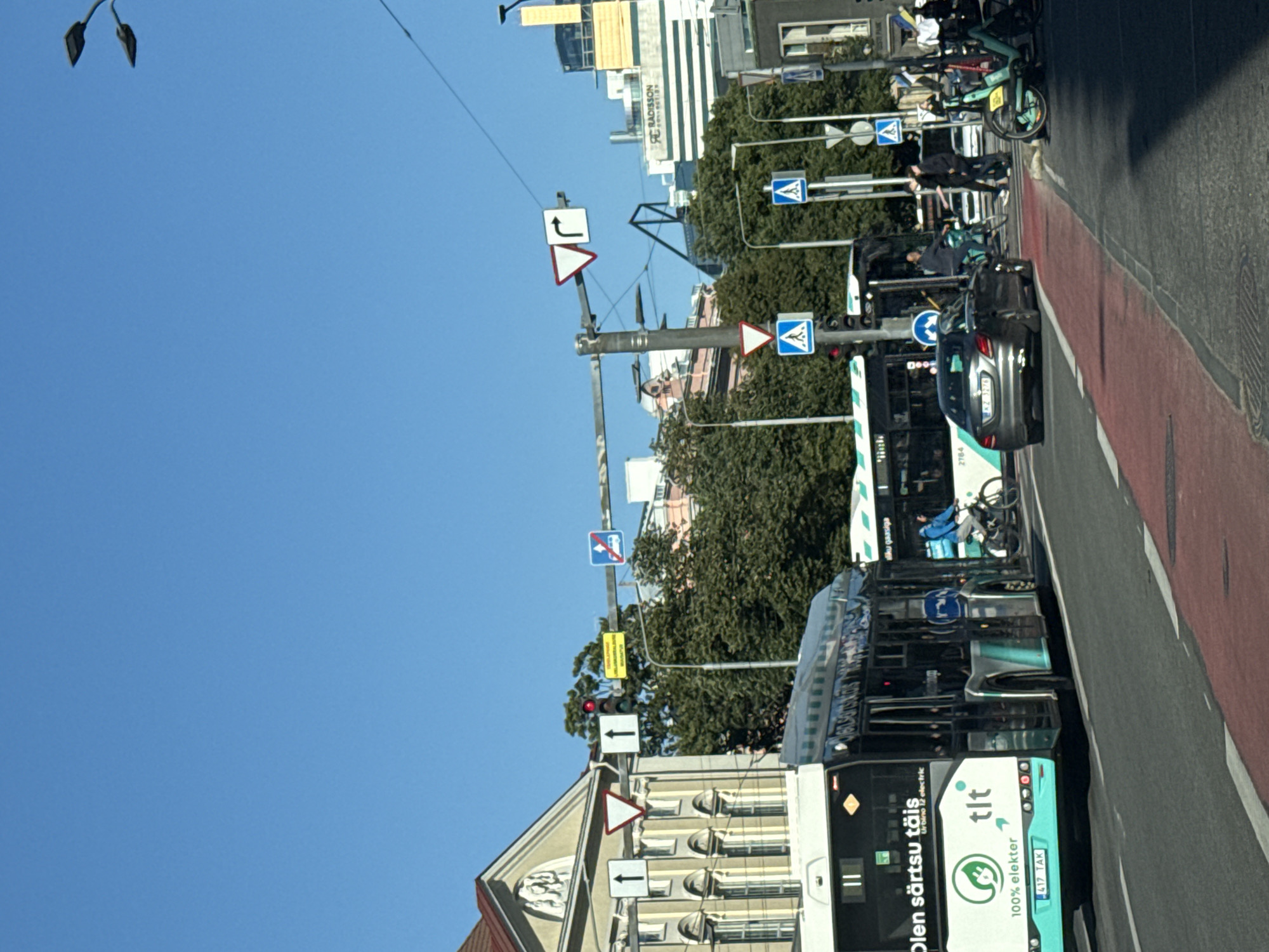 The image depicts a street scene in Tallinn, Estonia, on a clear, sunny day. In the foreground, a modern teal and white electric bus, displaying "TLT 100% elekter," is stopped at an intersection. To the right of the bus, a dark gray sedan, license plate "KZ 3276," is also at the intersection. Behind the bus, another bus is partially visible. The street is marked with red and white lines, indicating a pedestrian crossing or bike lane.
Traffic lights and various street signs are visible, including a red traffic light and directional arrows. In the background, trees line the street, and several buildings are visible, including one with a sign that reads "RADISSON." People are present, including a cyclist on the right side of the image and several pedestrians. The overall impression is a busy urban environment.