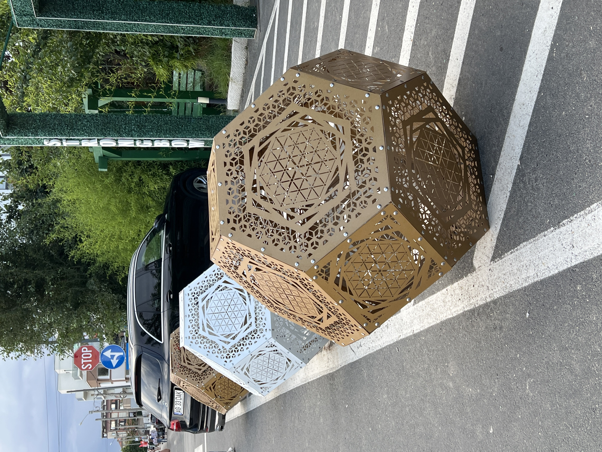This image captures a street scene in Năvodari, Romania, on a partly cloudy day. In the foreground, three large, geometrically shaped art installations are positioned along the edge of the road. The closest installation is a large, dodecahedron-like structure with intricate cut-out patterns in a metallic brown color. Behind it, a similar structure is visible in white, and another in a lighter brown. These installations appear to be placed as street furniture or artistic elements.
A black Mercedes-Benz SUV is parked to the left of the installations, with its license plate reading "AG 33 DAK". A white line marking a pedestrian crossing is visible on the asphalt. Further down the street, a red STOP sign is mounted above a blue directional sign indicating a left turn. Buildings with multiple floors line the street in the background, some with visible balconies. Lush green trees and vegetation are present on the right side of the frame, where a green structure with the word "STRONGBOW" vertically displayed on it is partially visible. This structure appears to be part of an outdoor seating area or an advertisement. Several people are walking in the distance on the left side of the street, but they are not clearly discernible. The overall scene suggests an urban environment with public art and a functioning street.