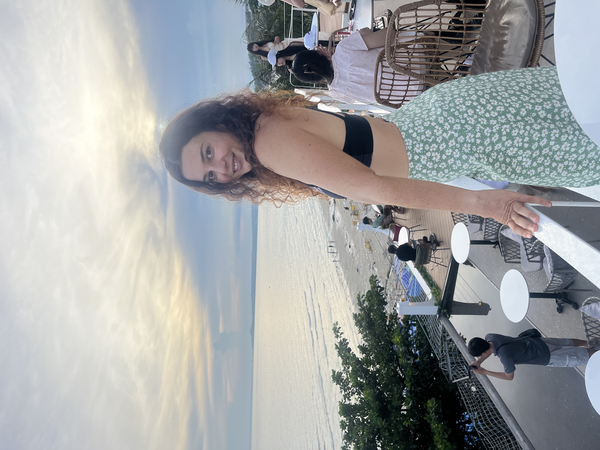 A woman with curly brown hair, wearing a black bikini top and a green and white floral skirt, stands on a balcony overlooking a beach and the ocean in Mũi Né, Vietnam. The sky is overcast with soft clouds, and the sun is setting, casting a warm glow over the scene. Several other people are visible on the balcony and on the beach, some sitting at tables and others walking along the sand. A man in a gray t-shirt and shorts is taking a photo with his phone. The balcony has white railings and tables with chairs. The overall atmosphere is relaxed and scenic.
