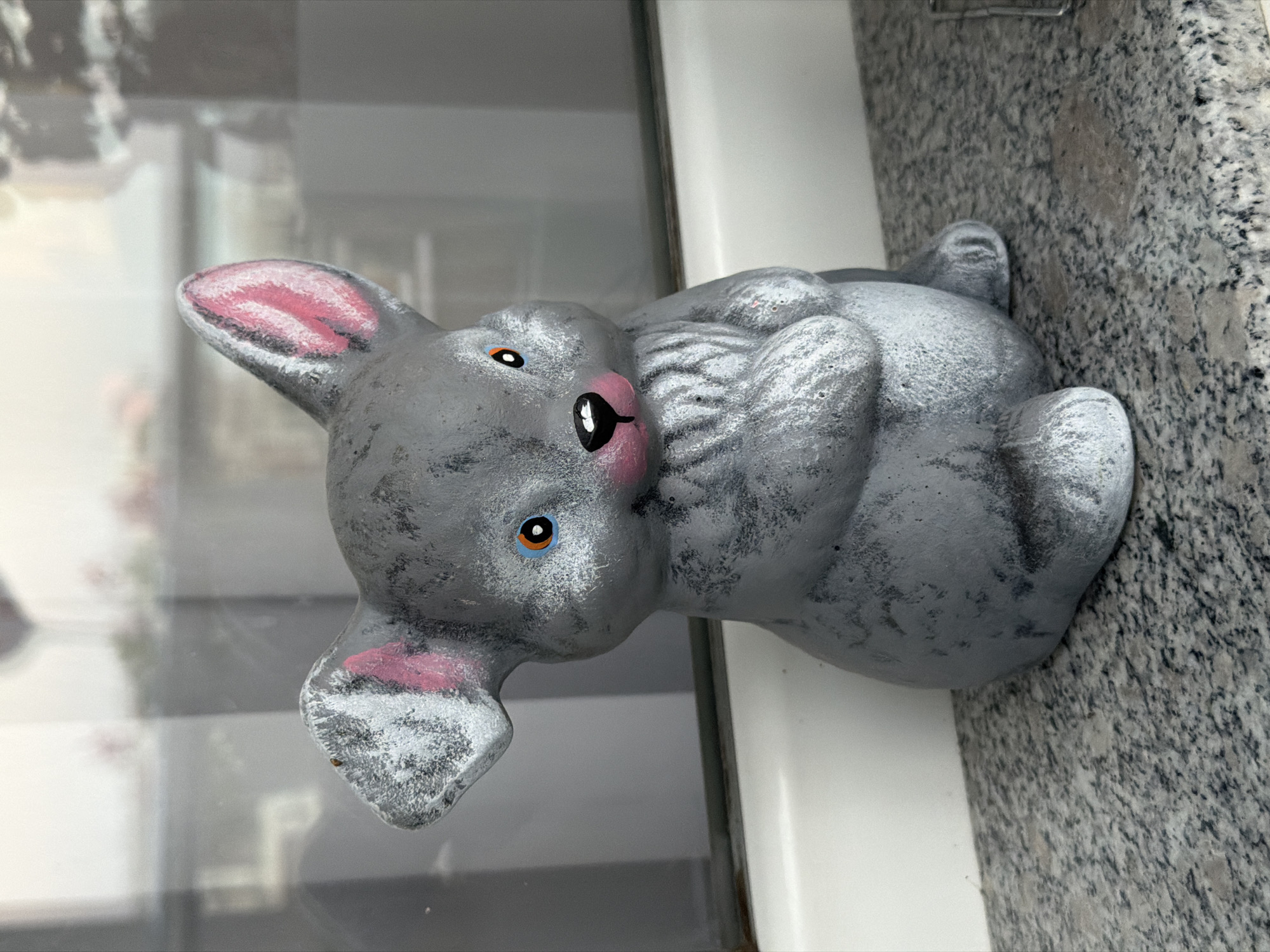 A grey ceramic figurine of a rabbit or mouse sits on a speckled grey granite countertop. The figurine has large ears with pink interiors, round orange and blue eyes, and a pink nose with a black outline. Its paws are held up in front of its chest. The figurine is positioned in front of a window with a reflection of trees and possibly furniture. A white ledge runs along the bottom of the window. The scene appears to be outdoors or in a room with a large window, possibly on a balcony or patio, in Sofia, Bulgaria.