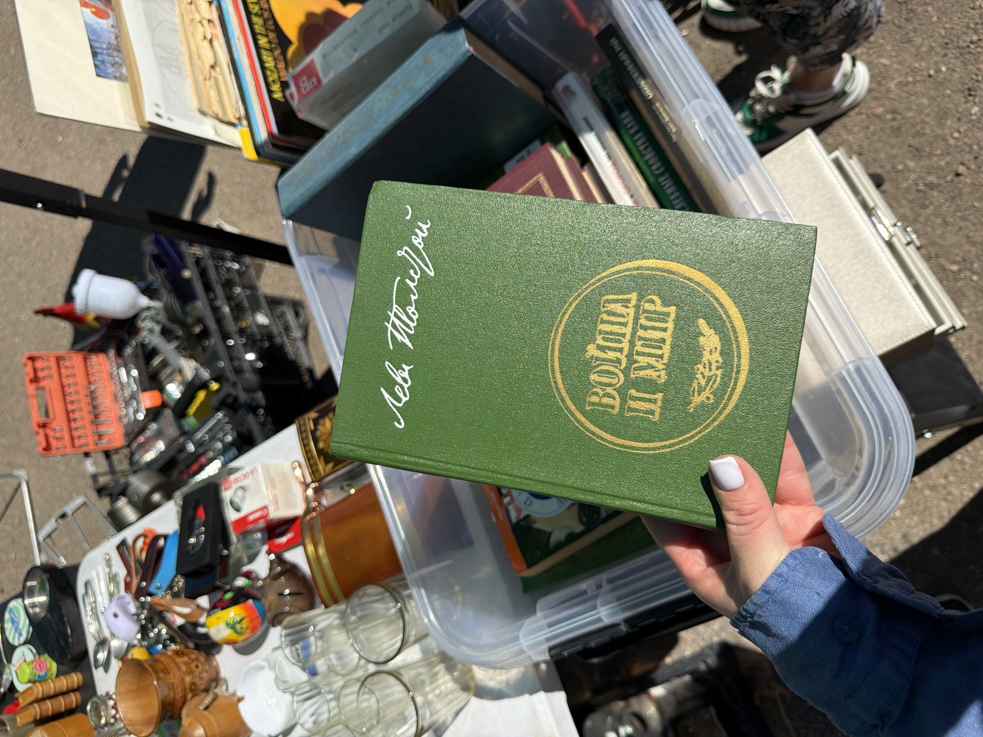 The image displays an outdoor market stall in Riga, Latvia, with a person's left hand holding a green hardcover book. The book features gold Cyrillic lettering, with "Лев Толстой" (Lev Tolstoy) at the top and "ВОЙНА И МИР" (War and Peace) enclosed in a circular emblem with a decorative branch motif below. The hand has light-colored nail polish on the visible fingers and wears a blue denim-like long-sleeved shirt.
The book is held above a clear plastic storage bin, which contains other books. To the left, on a white table surface, various items are spread out for sale, including wooden carved cups, small decorative objects, a silver bowl, and an assortment of metal cutlery. Further left, multiple clear glass tumblers are visible.
In the upper right background, an orange toolbox filled with tools, such as wrenches and sockets, is present. Below this, stacks of vinyl records are visible, with one sleeve partially showing text like "MOZART IN THE SEV..." and "WALDO DE LOS RIOS". Another clear plastic bin, also containing books, is visible to the right of the central book. The ground beneath the stall is a paved surface, likely concrete or asphalt, under bright sunlight, which casts distinct shadows. The feet of a person, wearing green and white sneakers, are partially visible at the bottom right of the frame.