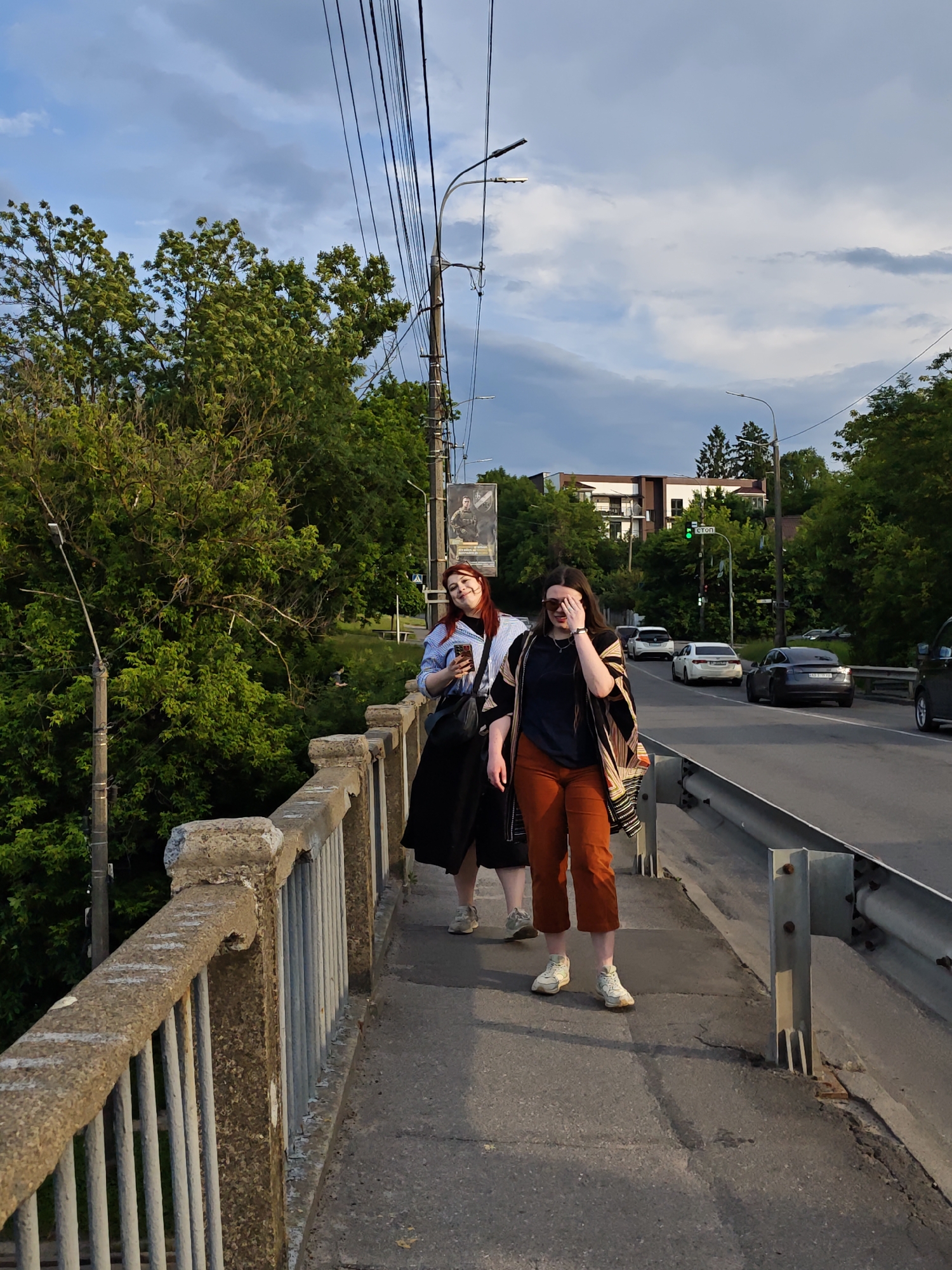 Two young women are walking on a pedestrian walkway beside a road. The woman on the left has red hair and is wearing a blue and white striped shirt and a black skirt. She is holding a phone in her hand and smiling. The woman on the right has dark hair and is wearing a dark t-shirt, orange pants, and a patterned shawl draped over her shoulders. She is covering her eyes with her hand.
The scene appears to be a bridge or overpass. A concrete railing with metal bars lines the walkway. Beyond the railing, a road with cars driving on it is visible. Trees and buildings are in the background. The sky is partly cloudy, suggesting it is late afternoon or early evening. A traffic light is visible in the distance, showing a green light. A poster is attached to a pole on the left side of the walkway.
The overall mood seems casual and relaxed, with the women seemingly enjoying a walk. The lighting suggests the sun is low in the sky, casting long shadows. The location cues like the cars and buildings suggest an urban or suburban setting in Vinnytsia, Ukraine. No discernible text is clearly readable on the poster or any other visible elements.
