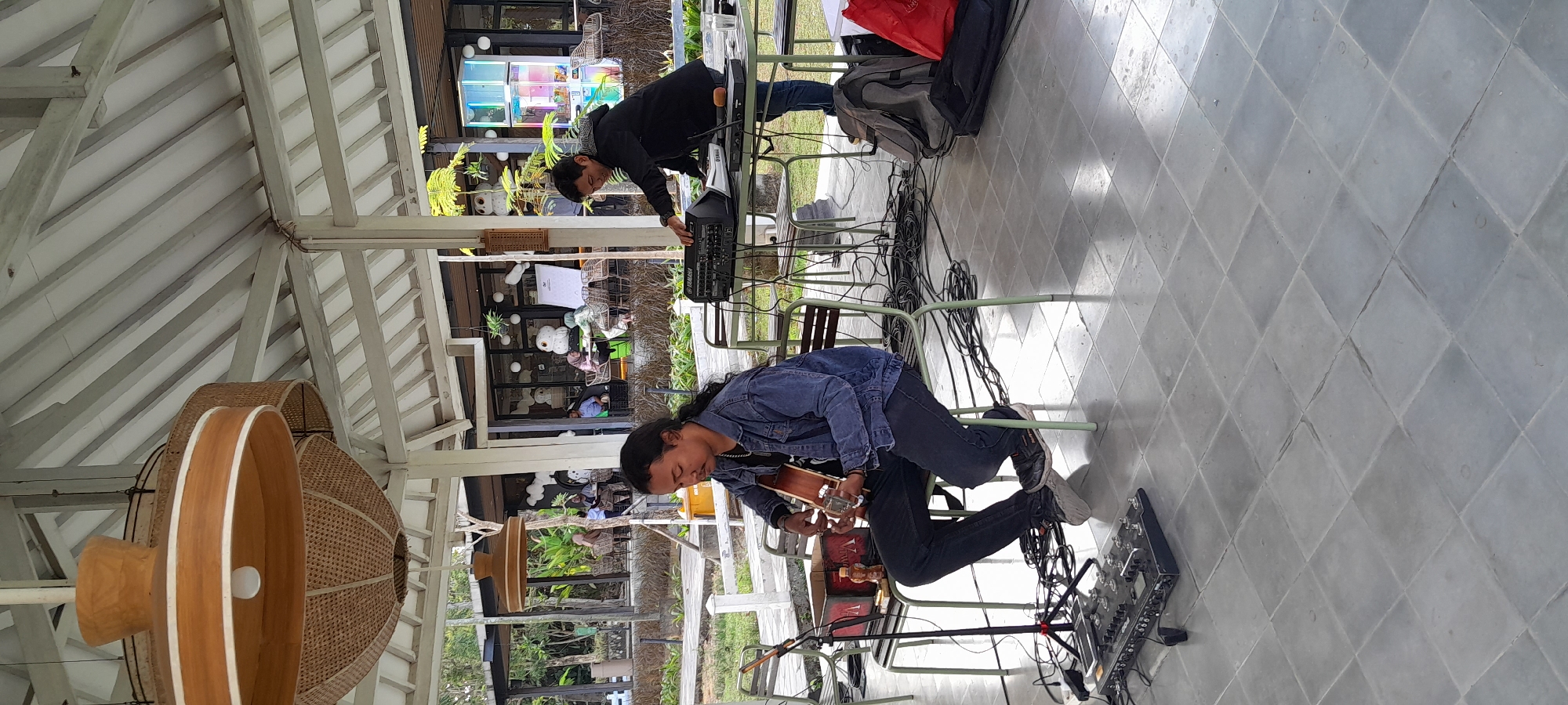 The image captures an outdoor performance scene at a venue located in Sleman, Indonesia. In the foreground, a man with long dark hair is seated on a green metal chair, playing an acoustic guitar. He is wearing a dark denim jacket and dark pants. In front of him on the tiled floor is a complex effect pedalboard and a microphone stand. To the right, another man is standing and appears to be operating an audio mixer or synthesizer on a table. He is wearing a dark hooded jacket and jeans. The background reveals a seated area with tables and chairs, where several people are visible, suggesting the venue is open to patrons. The architecture includes a white-framed roof structure with large woven pendant lights hanging from it. Greenery and trees are visible beyond the covered area. The overall environment suggests a relaxed, possibly casual, dining or entertainment setting.