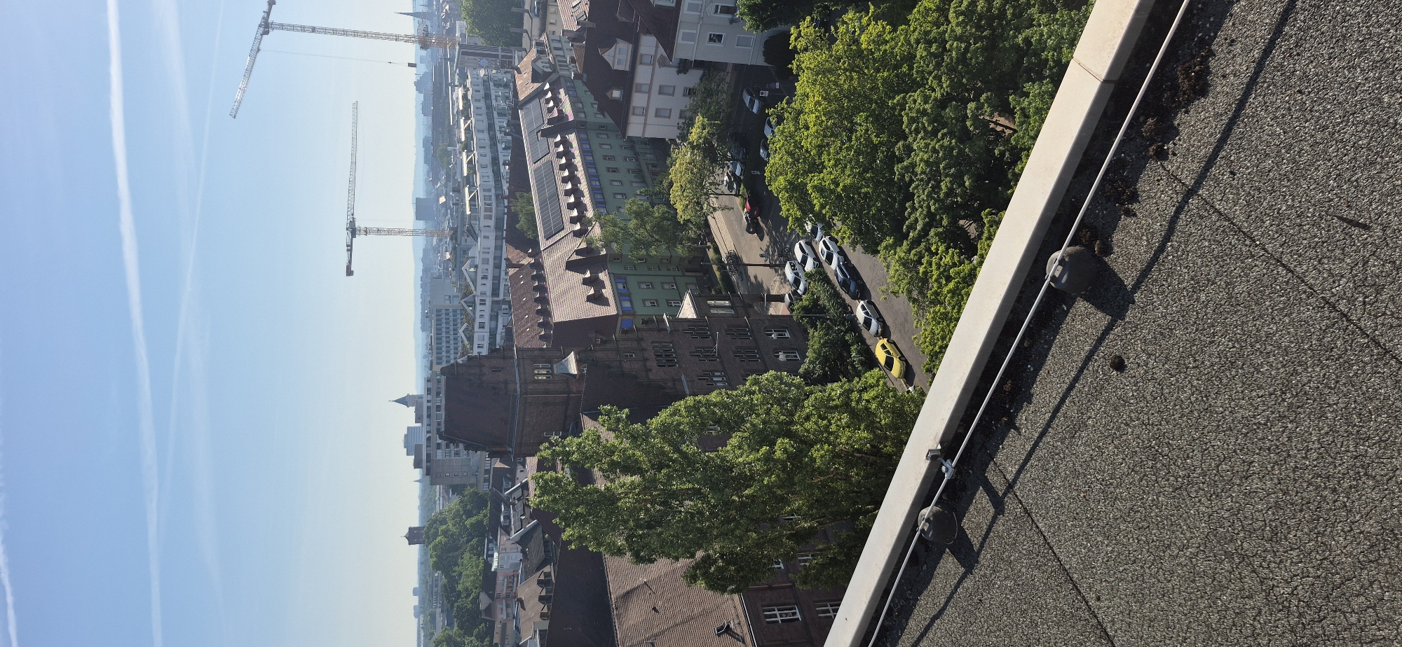 The image shows a cityscape with construction cranes visible in the distance. In the foreground, there is a rooftop with a grey, textured surface and some debris. Below the rooftop, a tree-lined street is visible with several parked cars. The buildings in the scene appear to be a mix of older, ornate structures and more modern buildings. The sky is clear and blue with some faint white clouds. Based on the distinctive red-tiled roofs and architectural style, this scene is likely located in the city of Karlsruhe, Germany, possibly in a residential or mixed-use area.