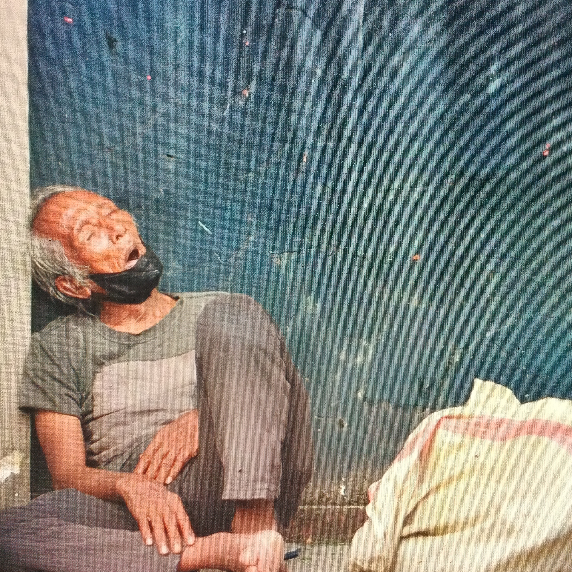 An elderly man with grey hair and a black face mask covering his mouth is slumped against a textured, dark blue wall, his head tilted back and eyes closed. He is wearing a muted green t-shirt over a lighter grey undershirt and loose-fitting grey pants. His legs are crossed, with one foot exposed. To his right, a large bundle of cloth, possibly a bag or blanket, is resting on the ground. The background wall has a rough, uneven surface with visible vertical lines and several small, reddish-orange specks. The lighting suggests an outdoor setting, possibly in a public urban space in Ende, Indonesia, given the context.