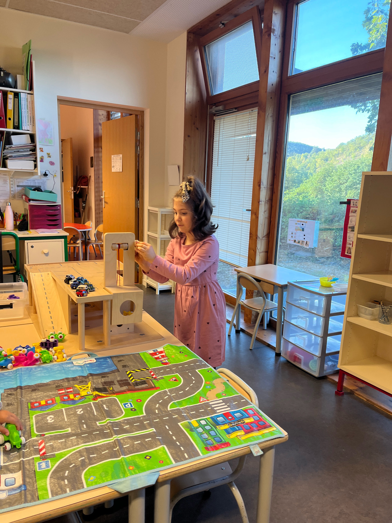 The image shows a young girl playing with a toy garage and cars in a room that appears to be a classroom or playroom. The room has large windows with a view of trees and hills, suggesting an outdoor setting in the city of La Fouillouse, France. The girl is wearing a pink dress and is interacting with the toy garage. There are other toys on the table, including a play mat with a road design and various small cars and figures. The room is well-lit, with natural light coming from the windows. Shelving units and a door are visible in the background, indicating a functional space.