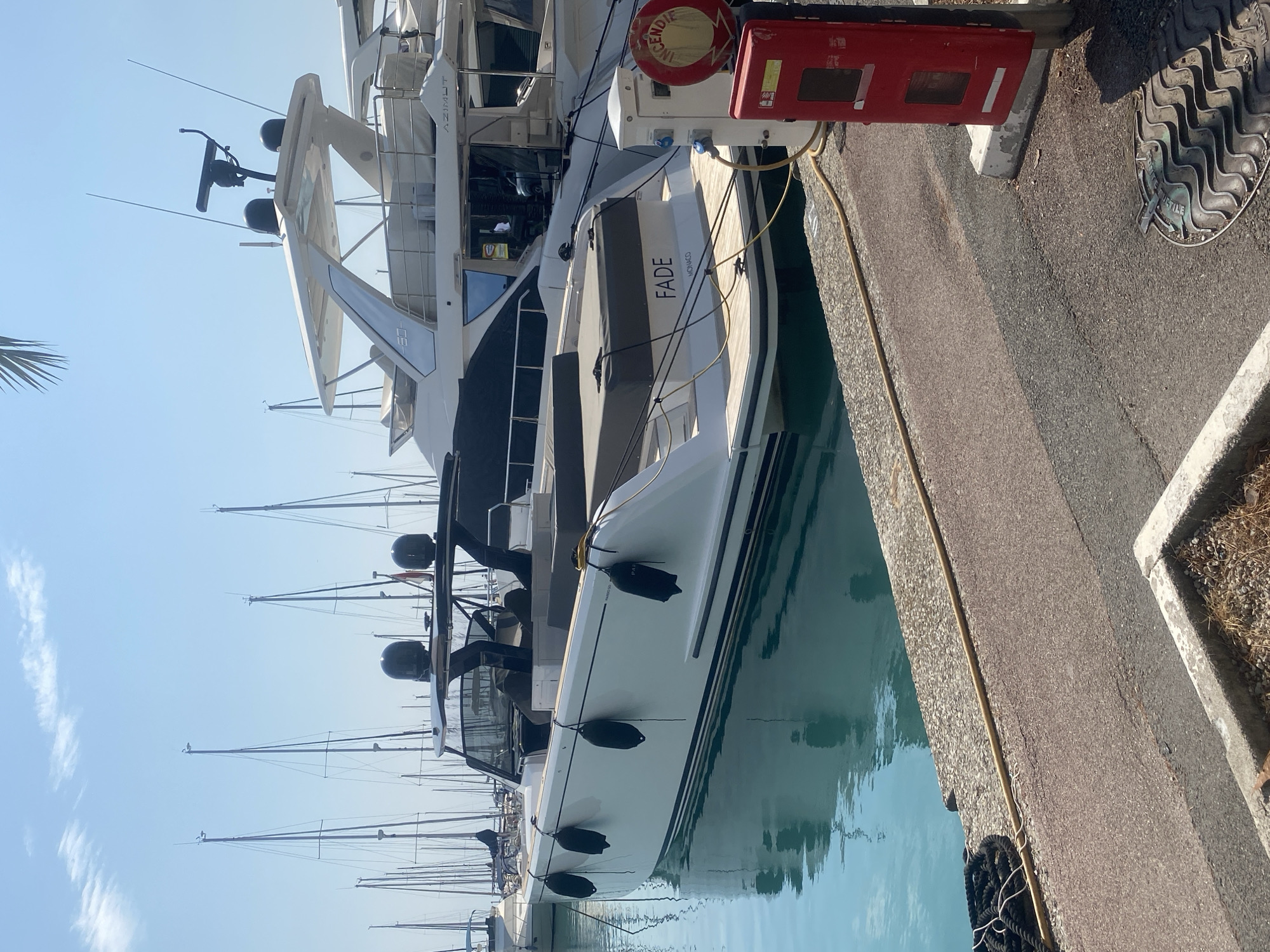 In Villeneuve-Loubet, France, a white yacht named "FADE" with a black canopy is docked in a marina. It is positioned in the foreground, with its reflection visible on the calm turquoise water. The yacht is connected to a power source by a yellow cable. Beside the yacht, a larger white yacht, identified as "AZIMUT," is also docked. A red fire safety box with an "INCENDIE" (fire) sign is situated on the dock next to the power source. In the background, numerous sailboats with tall masts are visible, along with a clear blue sky with scattered clouds. The dock is made of concrete and paved surfaces.