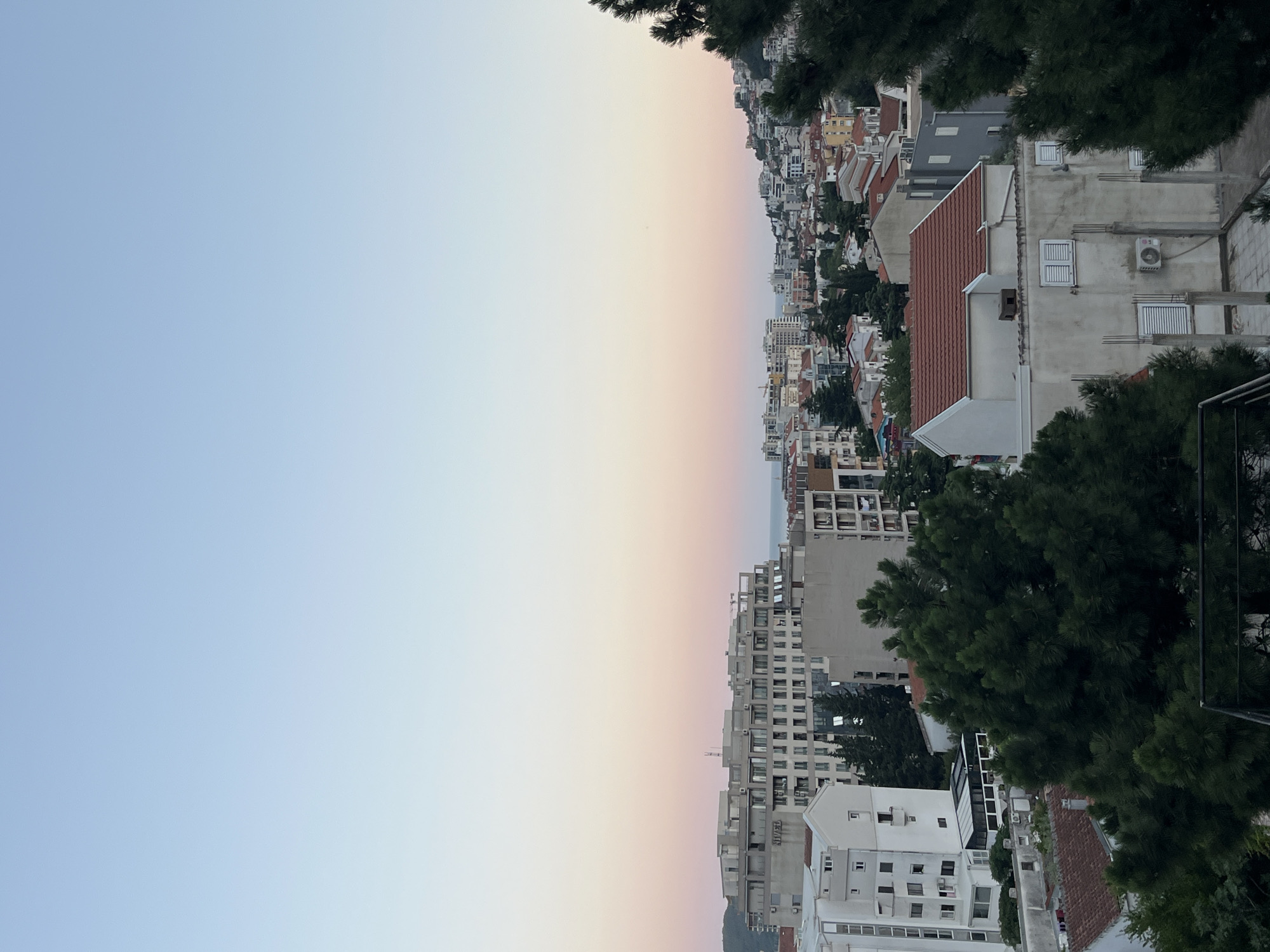 The image presents an elevated view of the cityscape in Budva, Montenegro, captured during conditions consistent with either sunrise or sunset. The sky occupies the upper half of the frame, displaying a clear gradient from a pale cerulean blue at the top to a soft pink and light orange immediately above the horizon. A thin, horizontal line, indicative of the sea, is visible on the distant horizon, extending across the central portion of the image.
The lower half of the composition is dominated by a dense urban environment interspersed with vegetation. In the foreground, dark green evergreen trees, likely conifers, partially obscure the immediate structures below; a dark railing or barrier is visible at the very bottom edge.
A prominent, multi-story building, light grey or white in color with numerous windows and balconies, stands in the midground, slightly to the left. The word "NIVEL" is clearly visible on its facade. A construction crane is partially discernible behind this building to the left.
The cityscape extends across the midground and into the background, characterized by a mix of building types. Predominantly, structures are white, grey, or light-colored concrete, frequently topped with red terracotta tile roofs. Various architectural elements are present, including air conditioning units, windows, some with closed shutters, and occasional laundry draped on balconies. The urban development continues onto the hills in the background, particularly on the right side, where buildings ascend the terrain. No individuals are visible within the scene.