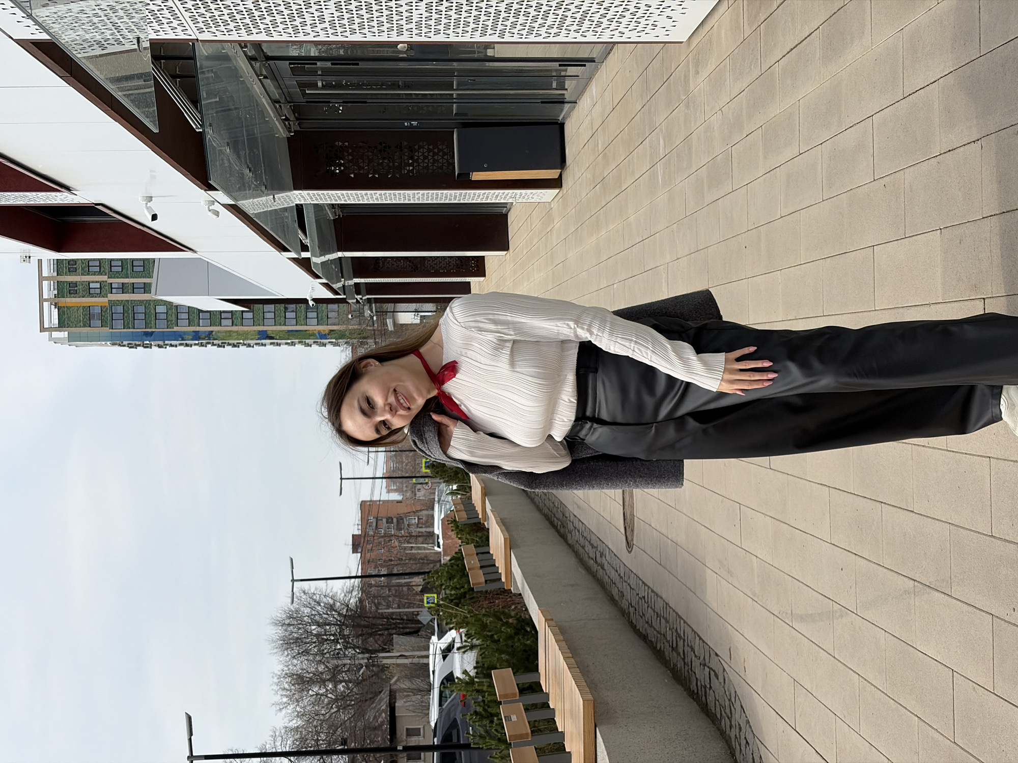 A medium, full shot depicts a woman with long brown hair standing outdoors next to a row of wooden benches on a paved sidewalk. The woman is angled slightly to the left, facing the camera with a smile. She wears a white, long-sleeved shirt, black pants, a red scarf, and a gray jacket draped over her shoulder. Her hands are resting on her hip.
Behind her, a street with parked cars and buildings stretches into the distance under an overcast sky. To the left, there is a modern building with geometric patterns on its facade. The photograph appears to have been taken in Izhevsk, Russia, as suggested by the context.