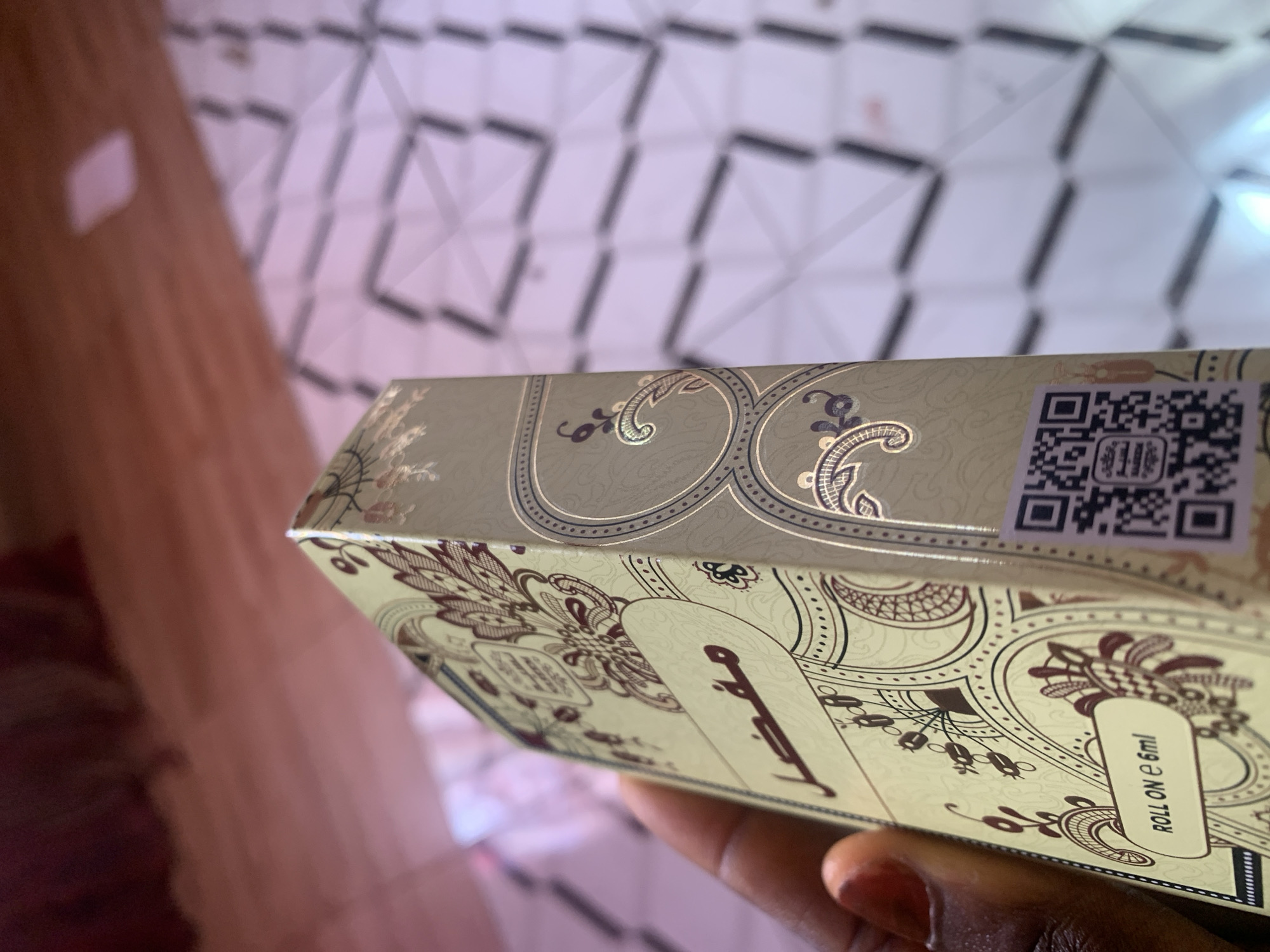 A hand, with dark skin and red-painted fingernails, holds a small, rectangular bottle with an ornate, gold and maroon design. The bottle is positioned at an angle, with the side facing the camera. Visible text on the bottle includes "ROLL ON e 6ml" and Arabic script. A QR code is also present on the side of the bottle. The background is blurred, showing a surface with a geometric pattern, possibly a tiled floor or ceiling, with a pinkish hue. The overall lighting is soft. The location is identified as Jauro Yinu, Nigeria.