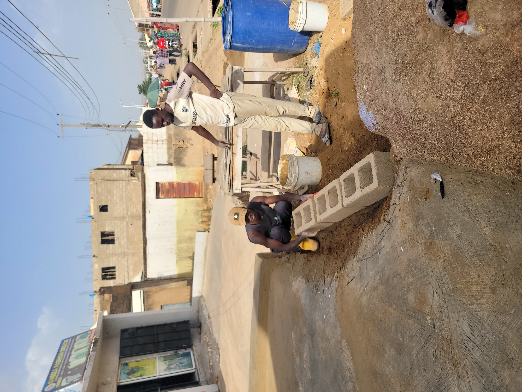 This image captures an outdoor scene in what appears to be an urban or semi-urban area, likely in Jos, Nigeria, during bright daylight hours under a clear blue sky. The setting suggests a mix of residential and commercial structures, with ongoing construction or development.
Two men are the primary subjects. On the right, a man stands with his hands on his hips, wearing a light-colored t-shirt with "MRV" and "BETANO" printed on it, paired with light-colored trousers. He looks towards the viewer. To his left and slightly in the foreground, another man is crouched, wearing a dark shirt, dark trousers, and a cap. He appears to be engaged in manual labor, possibly working with concrete hollow blocks which are visible near him, and is using yellow protective footwear or tools.
The immediate foreground features packed dirt and a concrete surface, with some exposed large rocks or boulders on the far right. A stack of concrete blocks is neatly arranged. Behind the standing man, a large blue barrel and two white plastic buckets are present, suggesting they hold water or building materials.
The background reveals a street or pathway lined with buildings in various stages of completion or repair. Some structures show exposed rebar and unpainted concrete blocks, indicating rudimentary construction. Overhead, multiple power lines crisscross against the sky, supported by utility poles. Further down the street, blurred figures, a green umbrella, and what might be market stalls or small shops are visible. A partial sign on a building in the distant left background reads "GUEEN FASHION DESIGN". The overall atmosphere is one of activity and everyday life in a developing urban environment.