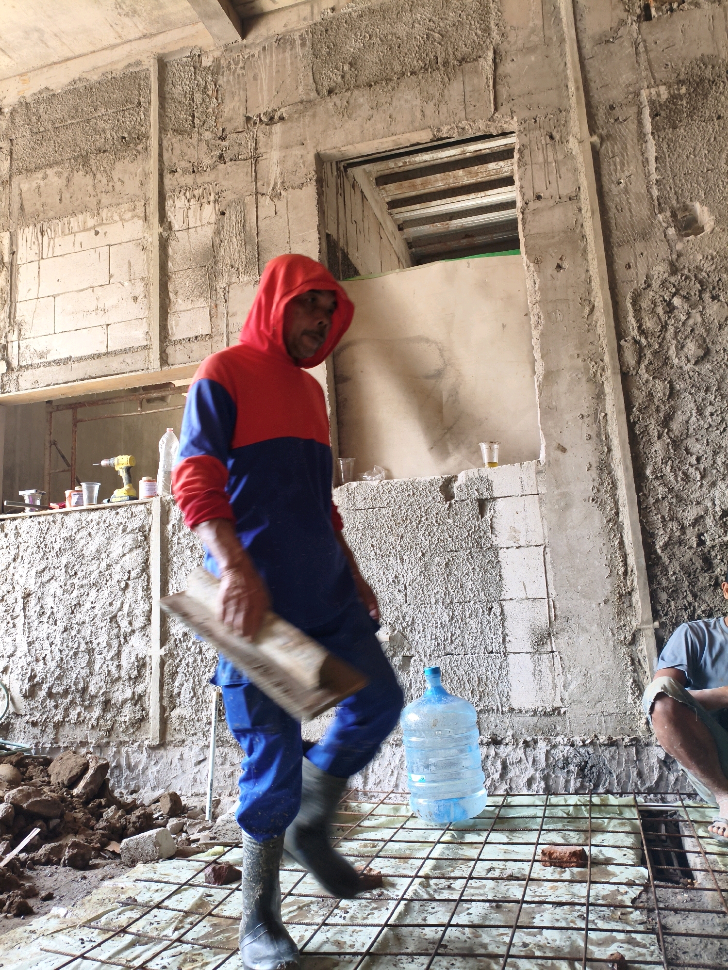 An outdoor, overhead shot shows a construction site in Bekasi, Indonesia. The scene is dominated by unfinished concrete walls and a floor covered in a wire mesh and plastic sheeting. A man, wearing a red and blue hooded sweatshirt and blue pants, walks across the mesh, holding a wooden plank. He is wearing dark boots. A large jug of water sits on the mesh near him. In the background, another person sits on the ground, wearing a light blue t-shirt and shorts. Construction debris, including piles of dirt and bricks, is scattered around the area. Tools, including a yellow drill, are visible on a shelf in the background. The lighting suggests it is daytime.