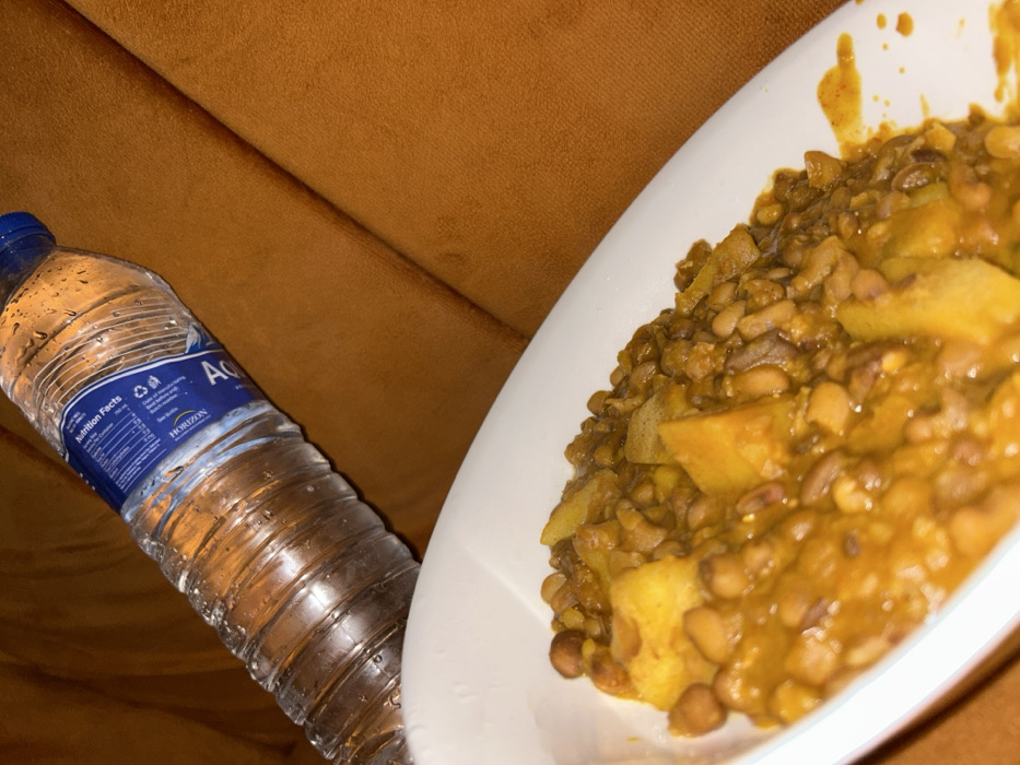 A water bottle labeled "Aqua" with a blue cap sits beside a white plate of cooked lentils and potatoes in a yellow curry sauce. The objects are set against a plush, orange surface, possibly a sofa or cushion. The water bottle is covered in condensation droplets. The food appears to be a hearty, home-cooked meal, likely a vegetarian dish.