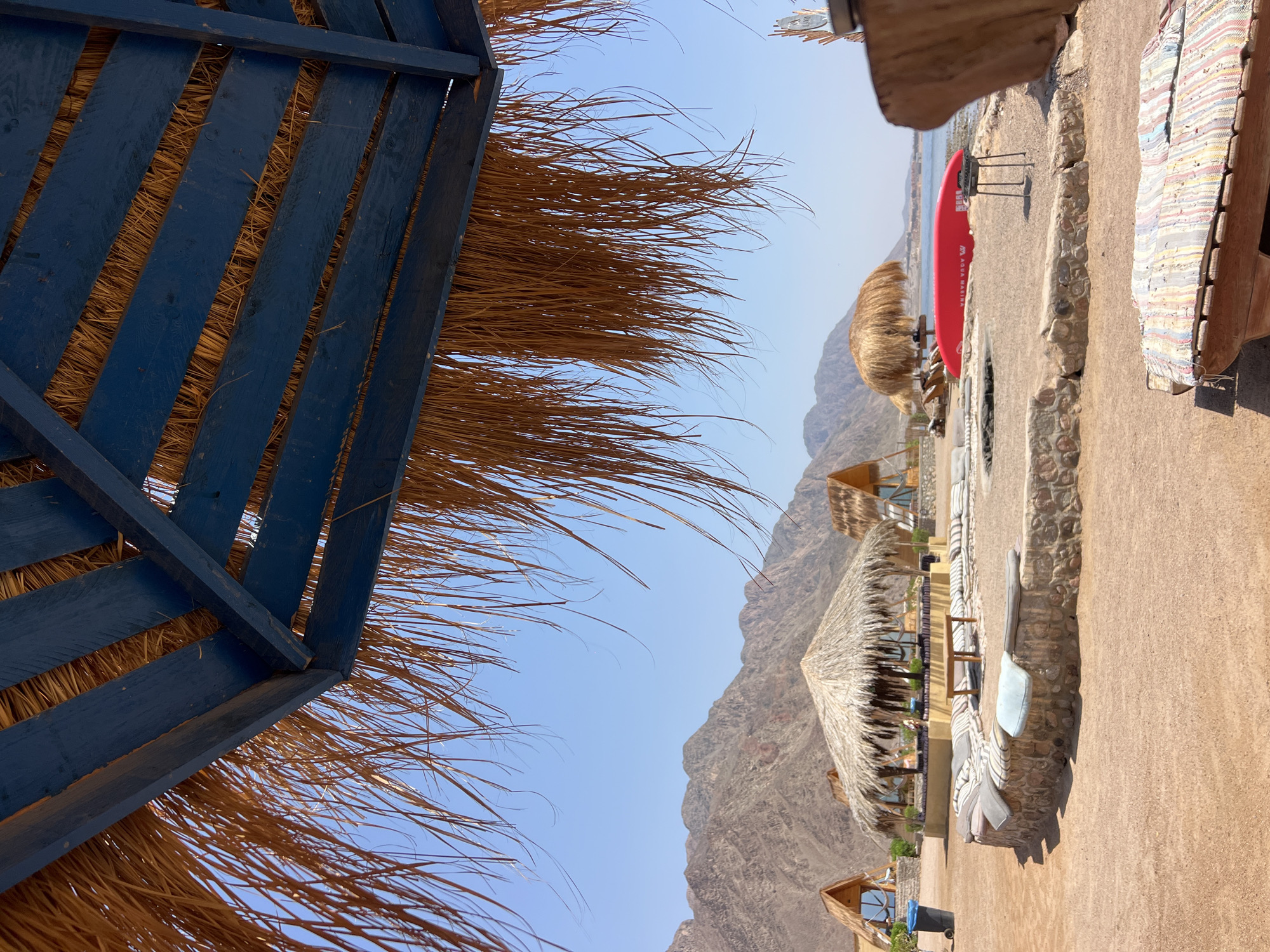 The image depicts an outdoor seating area at a resort in Nuweiba, Egypt, with a focus on the shaded lounging spaces. In the foreground, a thatch-roofed umbrella with blue wooden beams provides overhead cover, with dried palm fronds hanging down. Beyond this, the scene opens to a sandy area with several thatched-roof structures, resembling huts or cabanas, some with wooden frames and glass windows.
In the middle ground, a circular stone wall encloses a seating area furnished with numerous cushions. A small wooden table sits in the center of this area. Further back, another thatched-roof structure stands, this one more open, with seating arrangements visible beneath it. A bright red kayak rests on the sand near the water's edge, alongside a small barbecue grill. The background features the distinctive, arid mountain range typical of the Sinai Peninsula, under a clear blue sky. The presence of the beach, mountains, and resort-like structures strongly suggests a coastal location in Nuweiba, Egypt. No people are visible in the image.
