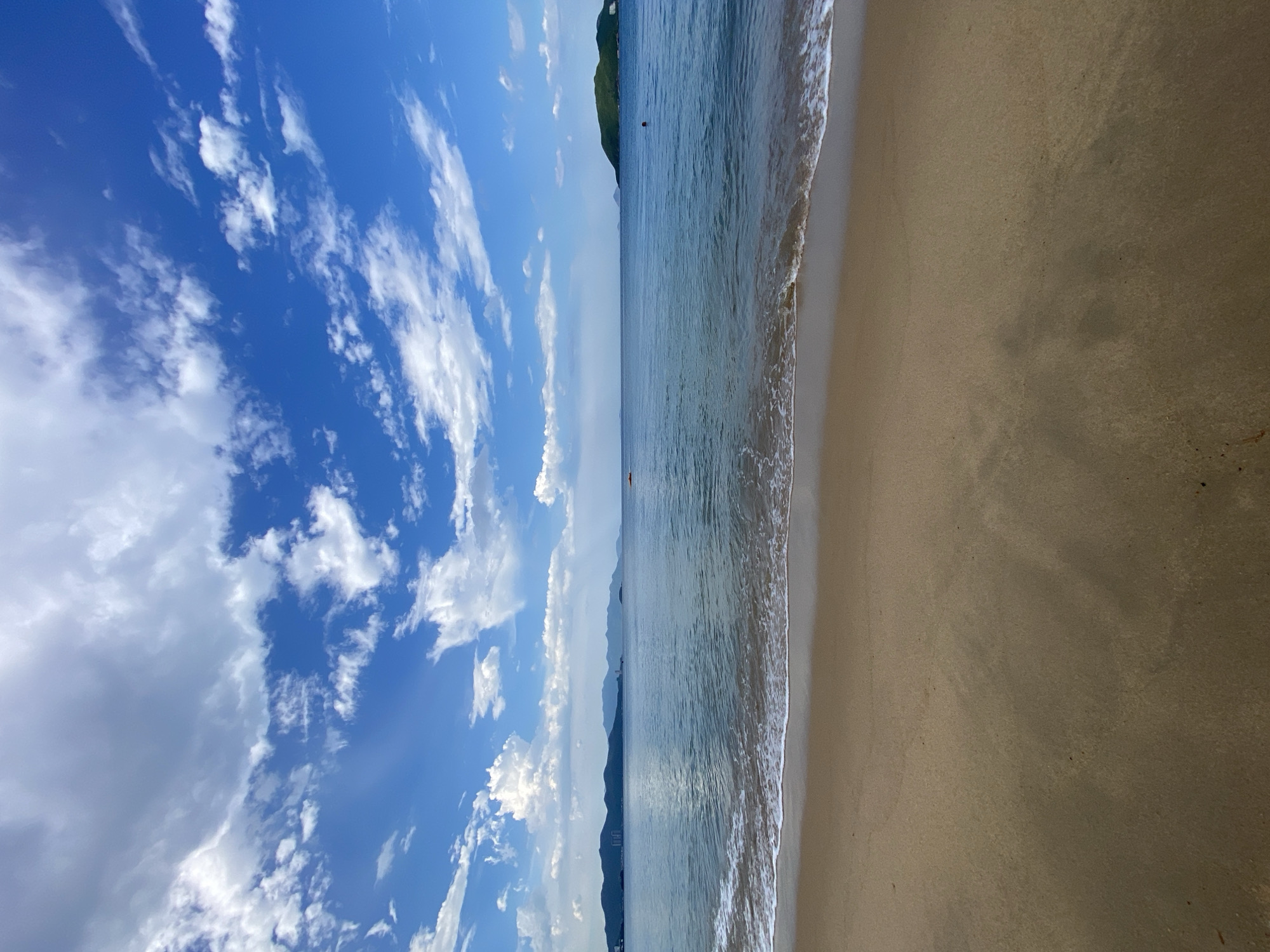 The image captures a serene beach scene in Nha Trang, Vietnam, likely during daylight hours given the bright sky. The foreground is dominated by a sandy beach, appearing damp and reflecting the light, with subtle shadows suggesting it's not mid-day. Gentle waves are seen lapping at the shore, creating a thin line of white foam.
Beyond the beach, the calm sea stretches out towards the horizon. In the distance, a chain of mountains forms the backdrop, with some appearing slightly hazy. Several small islands are also visible on the horizon, one of which is a larger, green, hilly island on the right side of the frame. The sky is a vibrant blue, adorned with scattered white clouds, some of which are fluffy and cumulus-like, while others are thinner and streaky. A faint hint of buildings can be seen along the distant coastline, suggesting proximity to urban areas.
There are very few signs of human activity. On the far right of the image, a single person is visible in the water, appearing as a dark silhouette. Further out in the water, towards the left, a small orange object, possibly a kayak or a boat, is also present. The overall impression is one of tranquility and natural beauty.
