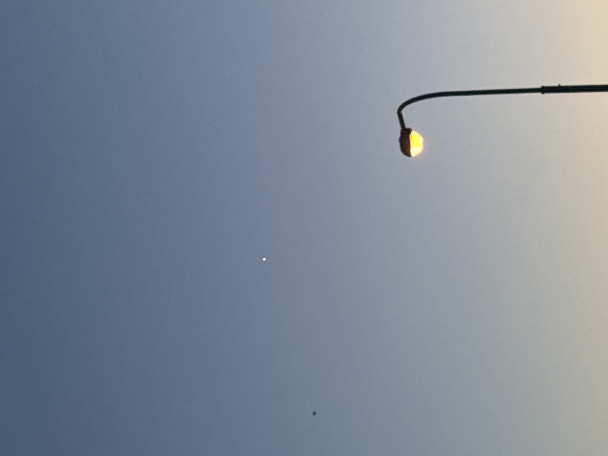 The image shows a clear sky with a gradient of blue, transitioning to a lighter hue towards the horizon. In the lower right portion of the frame, a street lamp with its yellow light on is prominently featured. In the upper center of the image, a small, bright white dot is visible, likely a star or planet. To the left of this celestial object, a smaller, dark circular object can be observed, which could be a bird or insect. The scene appears to be an outdoor setting in the city of Unknown, Unknown, at dusk or dawn.