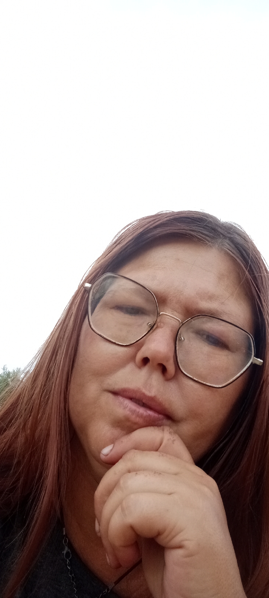 A low-angle, close-up photograph captures a person from the chest up, with their head tilted back and facing the camera. They are wearing oversized, angular eyeglasses with gold-toned frames. Their hair is a medium to dark reddish-brown color and falls past their shoulders. The person's right hand is raised, with their chin resting on their index finger and thumb, and the other fingers curled inward. They are wearing a dark gray garment, possibly a shirt or sweater, and a necklace is visible. The background is a bright, overcast sky. The scene appears to be outdoors in a natural setting, as indicated by a faint hint of greenery on the far left edge of the image, suggesting a rural environment. The location of Январцев ауылдық округі, Kazakhstan, is not discernible from the visual elements of the photograph itself.