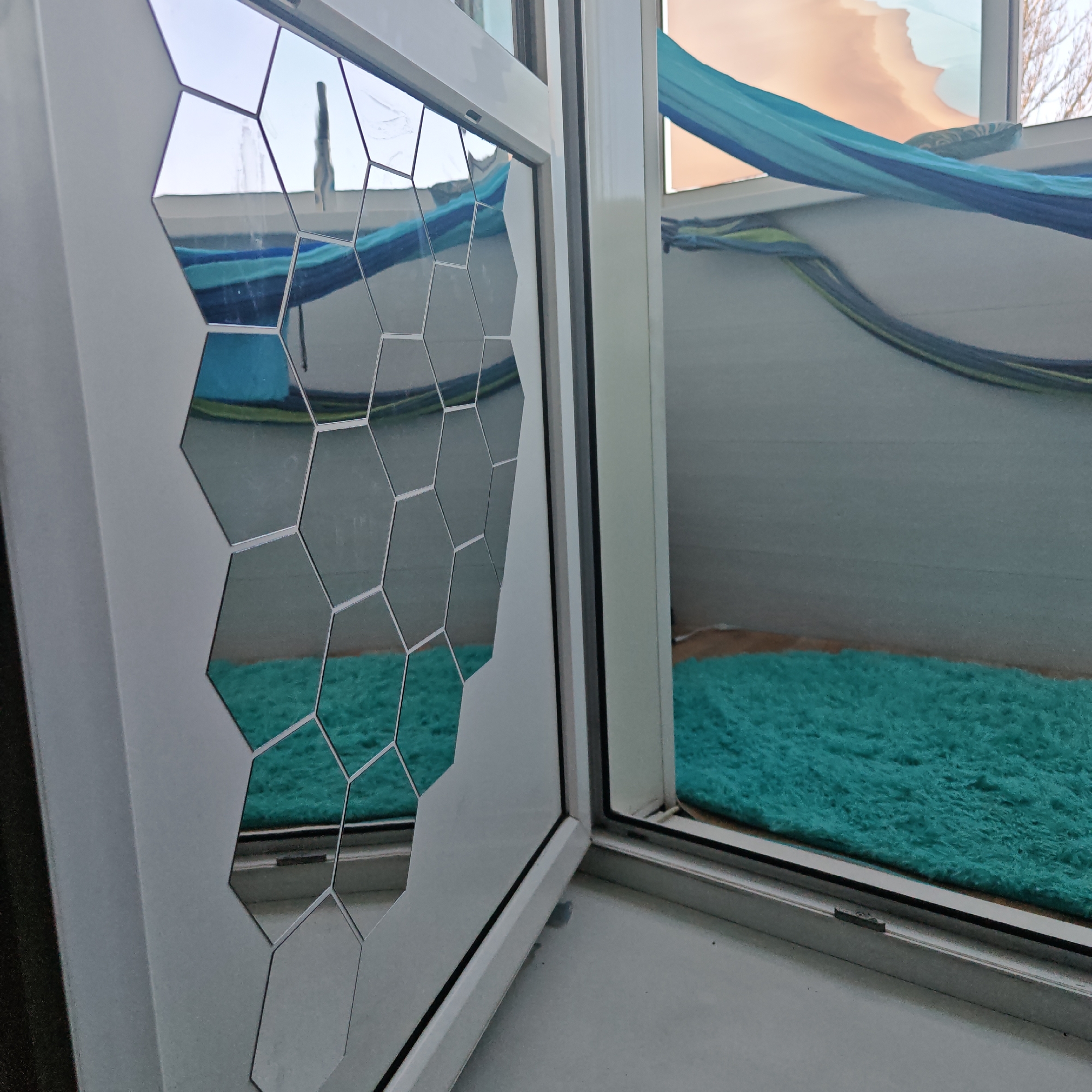 The image presents an interior perspective looking through a partially opened white window, situated in Rostov, Russia. The interior face of the left window pane is decorated with a pattern of hexagonal mirror tiles, which reflect elements of the scene. These reflections include a blue and light-blue striped fabric hammock, a teal-colored shag rug, and a distant cityscape containing a tall, dark, slender structure and a fountain displaying visible water spray, all under a bright sky. Beyond the open window on the right, the same blue and light-blue striped hammock is visible, suspended over what appears to be an exterior balcony or terrace. The floor of this exterior area is partially covered by the teal shag rug. In the upper right section of the main window, an orange-toned sky and a tree with bare branches are discernible. The window sill is light gray.