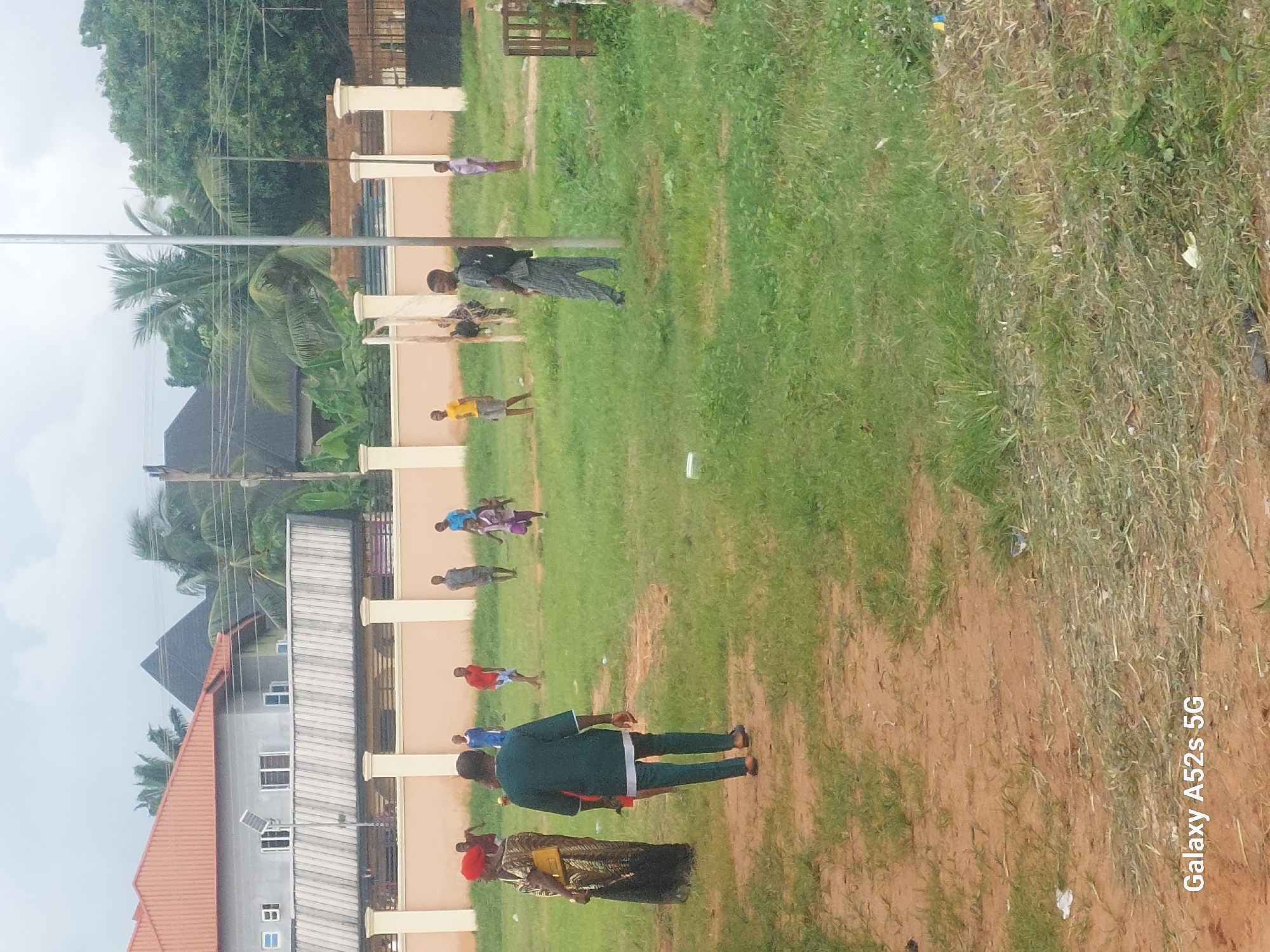 The image depicts an outdoor scene in Umudioka, Nigeria, featuring a group of people on a grassy field. In the foreground, two individuals are prominent: a woman in a colorful patterned dress and headwrap, and a man in a dark green traditional outfit. The man is walking away from the camera. In the background, several children and young adults are scattered across the field. Some appear to be playing or interacting with each other. Buildings with distinctive orange and grey roofs are visible in the distance, along with palm trees and other greenery, characteristic of the Nigerian landscape. A light pole stands near the center of the image. The ground is a mixture of grass and dirt paths. The overall atmosphere suggests a casual gathering or activity taking place in a residential area.