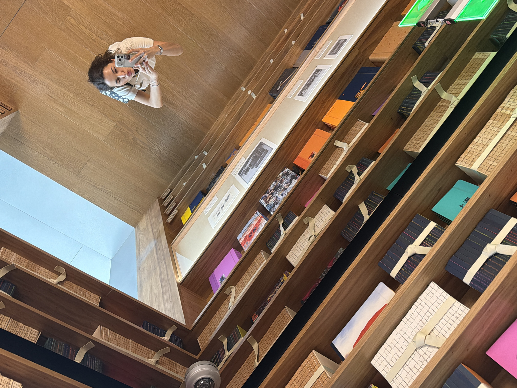 In Jing'an District, China, a woman holds a smartphone, facing the camera, presumably taking a selfie. The setting is a wooden-paneled building with shelves stocked with various merchandise. The building has a high ceiling with a skylight. The woman is wearing a light-colored top and has dark hair. The shelves contain items, some of which are adorned with ribbons.