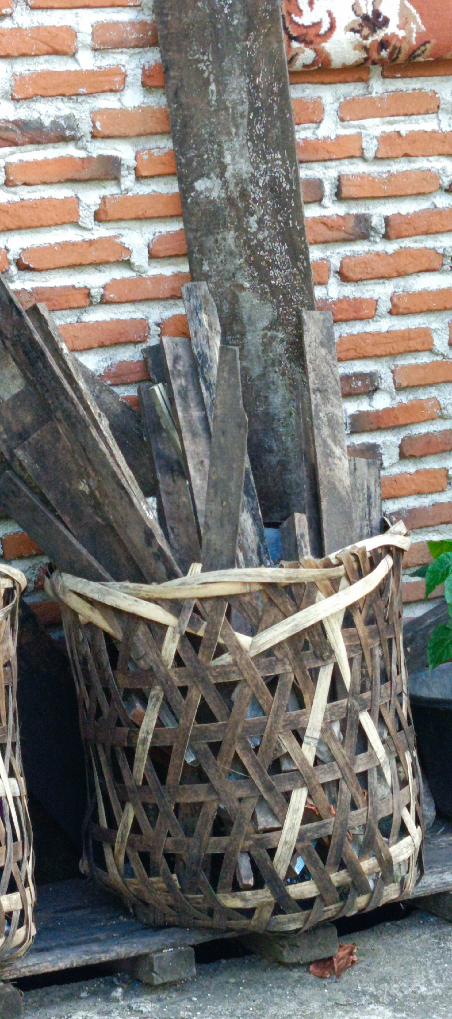 In Pashiebeutong, Indonesia, a woven bamboo basket filled with a collection of weathered wooden planks is positioned in front of a textured red brick wall. The basket's latticework, constructed from both light and dark bamboo strips, creates an intricate pattern of geometric openings. The wooden planks are of varying sizes and conditions, some appearing rough and splintered, others smoother and darker with age. They are stacked haphazardly within the basket, with some extending above its rim. A large, dark, flat piece of material, possibly slate or aged wood, stands behind the planks, leaning against the brick wall. The brick wall itself is composed of orange-red bricks with visible mortar joints, some of which appear to be crumbling. A small section of a patterned textile, featuring earthy tones and floral motifs, is visible at the very top of the image, resting on the brick wall. The ground beneath the basket is a rough, grayish surface, with scattered small stones and debris. The overall scene suggests an outdoor storage area, possibly for building materials or salvaged items.