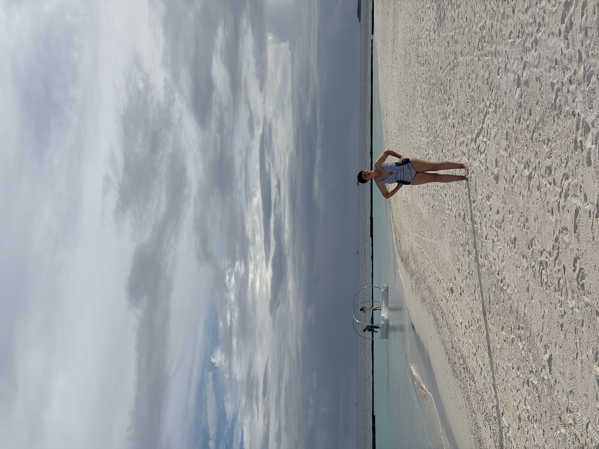 The image depicts a woman standing on a sandy beach in Halaveli, Maldives. She is wearing a blue and white striped one-piece swimsuit and has her hands on her hips, looking towards the camera. The beach is made of light-colored sand with some footprints and tire tracks visible. The water is a clear turquoise, gently lapping at the shore. In the distance, there is a white circular structure floating on the water with two people standing on it. The sky is overcast with dramatic, dark clouds. The scene appears to be set during the day, with diffused light due to the cloud cover.