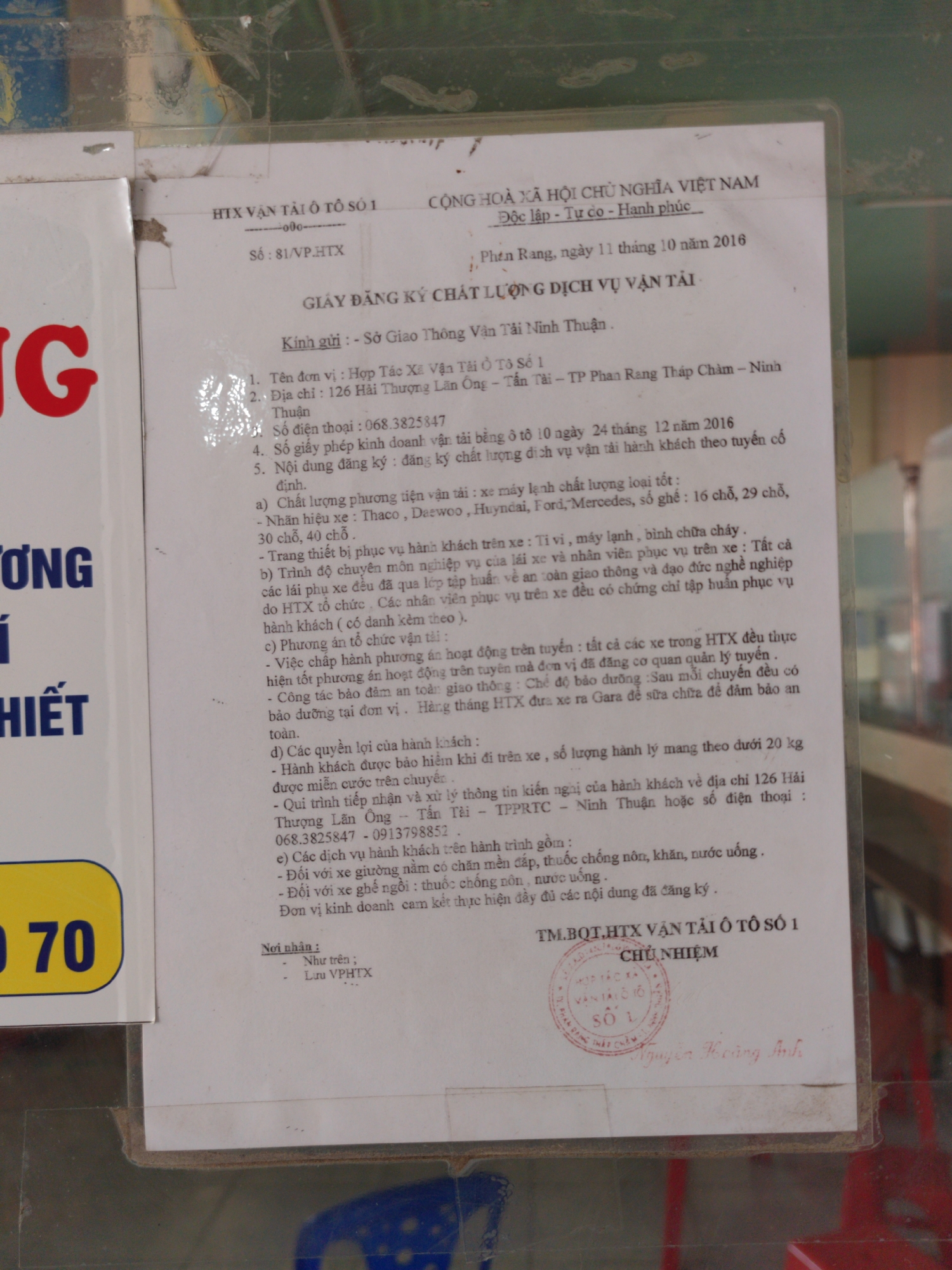 A framed document titled “GIẤY ĐĂNG KÝ CHẤT LƯỢNG DỊCH VỤ VẬN TẢI” (QUALITY REGISTRATION OF TRANSPORTATION SERVICES) is affixed to a surface. The document is addressed to Sở Giao Thông Vận Tải Ninh Thuận (Department of Transport of Ninh Thuận). The document provides details about Hợp Tác Xã Vận Tải Ô Tô Số 1 (Transportation Cooperative No. 1), including the location at 126 Hải Thượng Lãn Ông - Tấn Tài - TP Phan Rang Tháp Chàm, Ninh Thuận; phone number 068.3825847; transport license; vehicle quality; facilities; operation; customer rights; customer information, phone numbers, and services for passengers. The document has a red stamp and signature from Nguyễn Hoàng Anh.