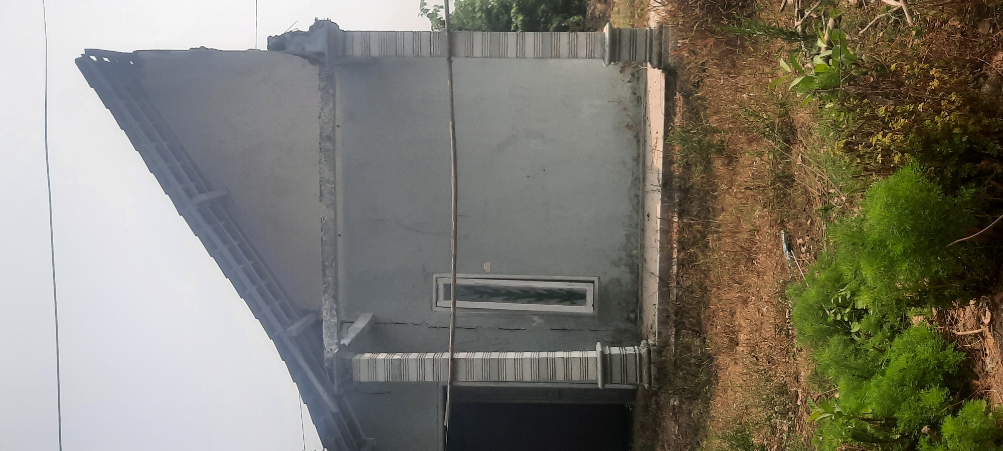The image shows a partially constructed or damaged house in Ploso, Indonesia. The house has a grey exterior with visible cracks in the plaster, particularly around the porch columns. There are two striped decorative columns supporting the porch roof. A narrow, vertically oriented window with a green decorative pattern is visible on the front wall. A wooden pole or beam runs horizontally across the wall. The roof has exposed rafters and appears incomplete or damaged at the peak. The area around the house is overgrown with dry grass and some green bushy plants in the foreground. The sky is overcast and appears hazy. No people are visible in the image.