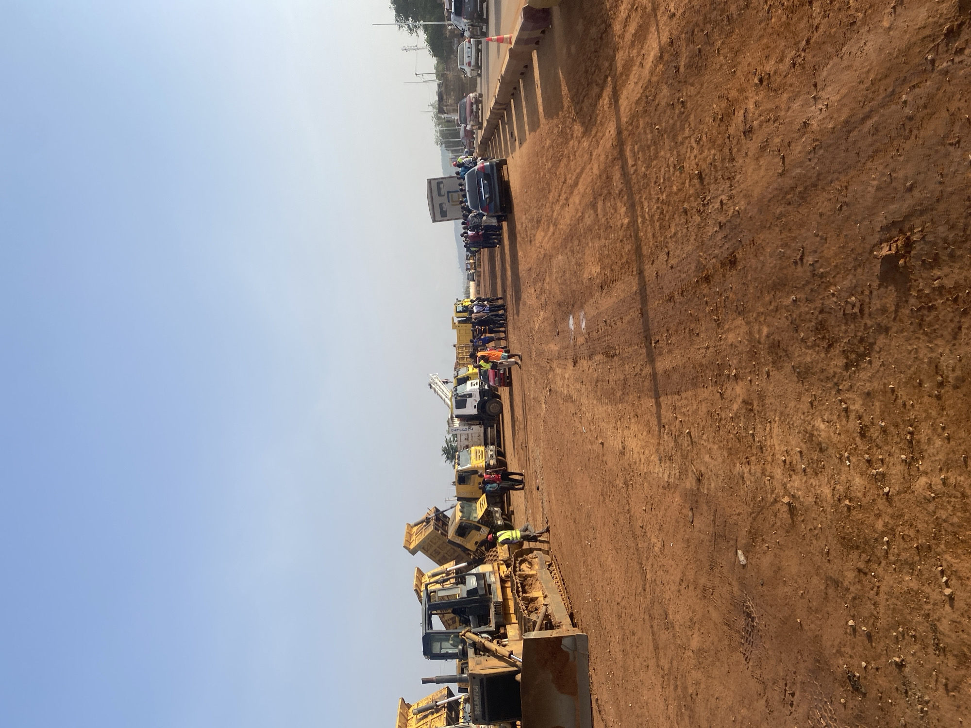 The image shows a construction site in Abuja, Nigeria. Heavy machinery, including bulldozers and trucks, are lined up along a dirt road. A group of people, likely construction workers, are standing near the machinery. A bridge or overpass is visible in the background, with vehicles and more people on it. The sky is clear and blue. The ground is reddish-brown dirt.
