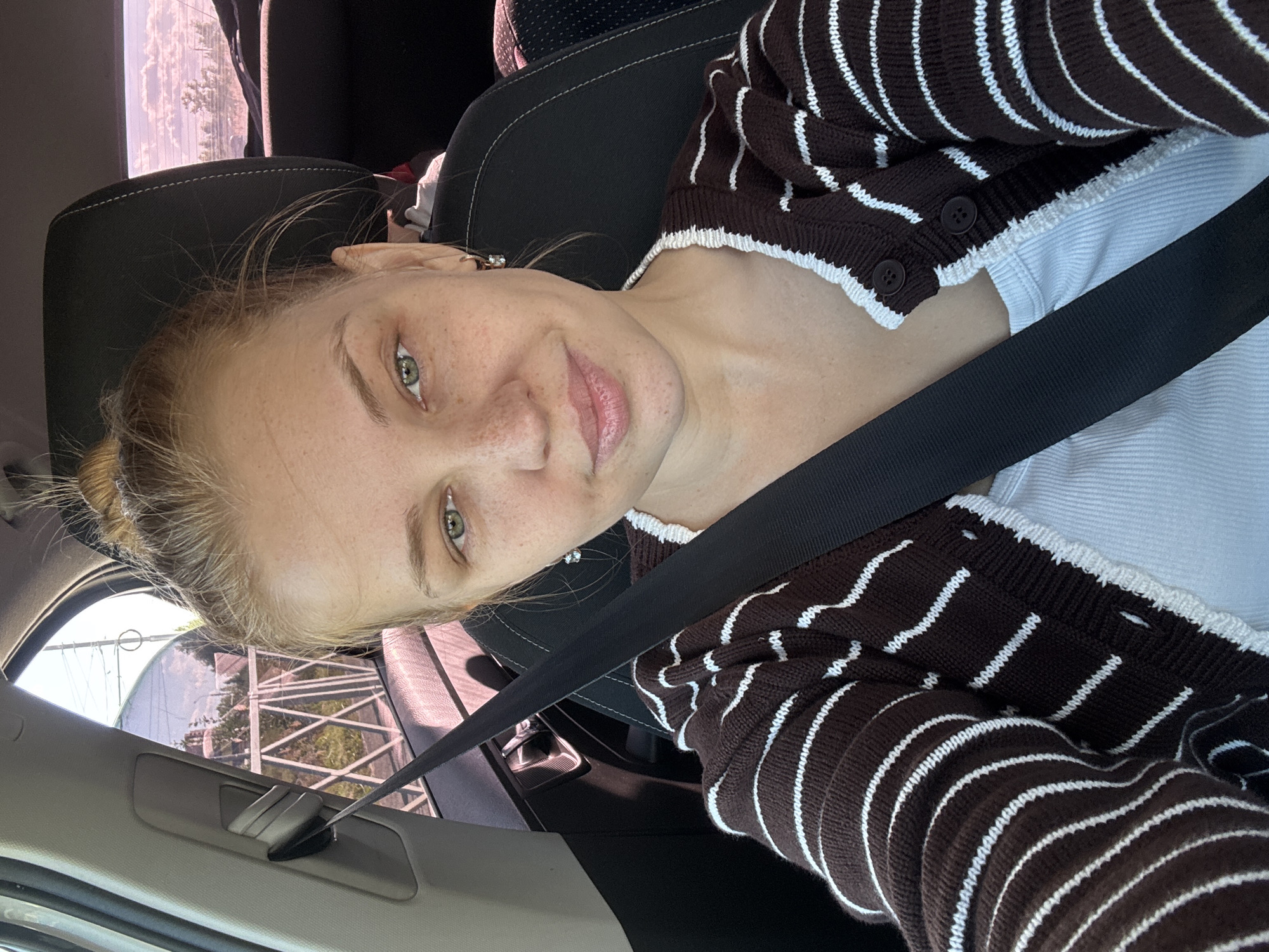 The image shows a young Caucasian woman sitting in the passenger seat of a car. She is wearing a white ribbed tank top and a brown and white striped cardigan. Her hair is pulled up in a messy bun. She has green eyes and freckles. The background shows a view from the car window of a street with trees and houses, suggesting she is in a residential area of the city of Rusanovo, Russia. A black seatbelt is visible across her chest.