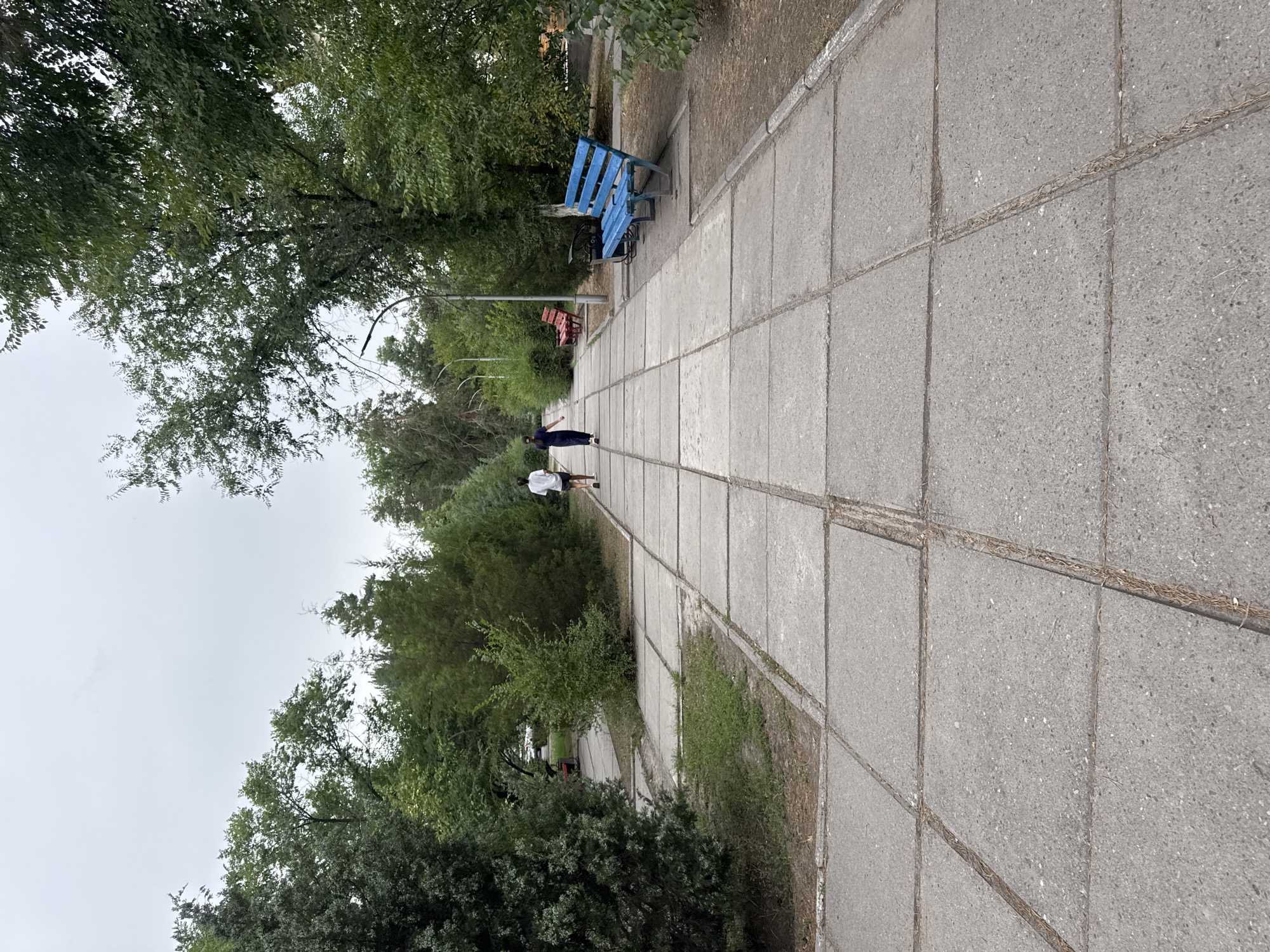 The image shows a park path in Kara-Balta, Kyrgyzstan. The path is made of large, grey concrete slabs. Two people are walking away from the camera on the path: one in a white t-shirt and dark shorts, and the other in a dark blue dress. To the right of the path, there is a bright blue park bench and a trash can. Further down the path, another red bench is visible. The path is lined with trees and bushes on both sides, and the sky is overcast.