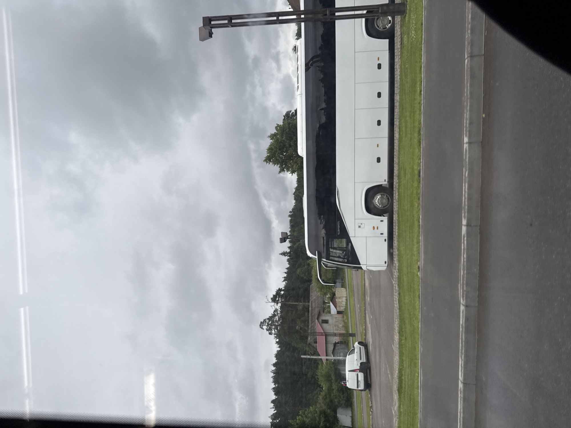 In the city of Rassudovo, Russia, a white bus with a black tinted window is parked in a lot. The bus is long and has multiple windows. In the background, there are trees and buildings. There is also a white van parked in the lot. The sky is cloudy and the ground is wet, suggesting that it may have rained recently. A lamppost is visible on the right side of the image.