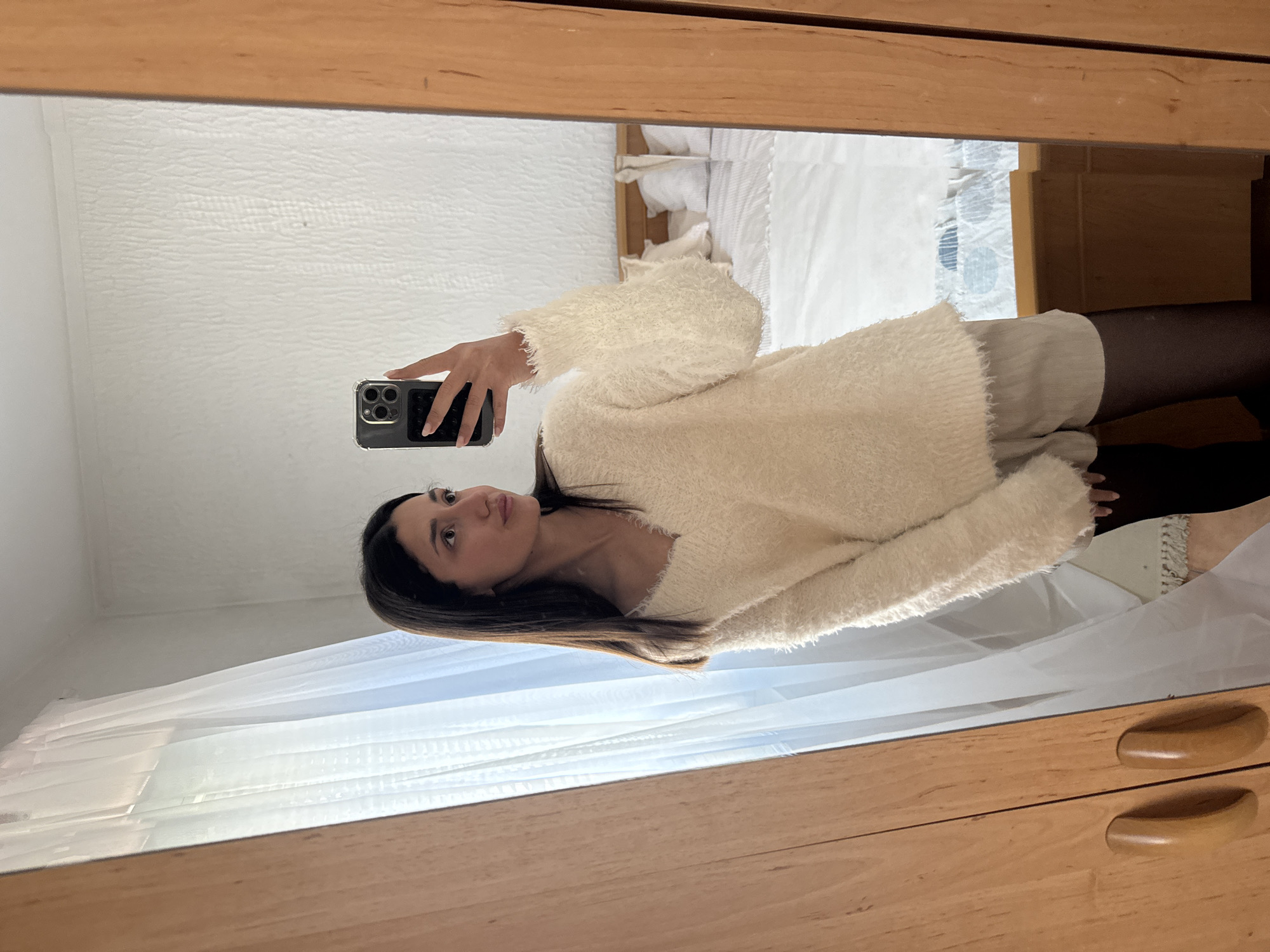 A mirror selfie captures a young adult female standing in an indoor setting, identified as located in Sutomore, Montenegro. The female subject, positioned centrally, has long, dark hair and is wearing a cream-colored, fluffy, V-neck long-sleeved sweater, light beige shorts, and dark opaque tights. She holds a dark-colored smartphone, equipped with a clear case and a black wallet attachment, in her raised right hand, pointed towards the mirror. Her head is turned slightly to her right, with her gaze directed to her right. The rectangular mirror features a light-brown wooden frame and reflects portions of the room. Visible in the reflection on the left is a window covered by sheer white curtains. Behind the subject, a bed with a light-colored wooden headboard and white bedding (including pillows and a duvet) is present. The wall behind the bed and subject is light-colored and features a discernible textured surface. A segment of a wooden wardrobe with two oval wooden handles is visible on the far left of the image. A low, light-brown wooden furniture piece is partially reflected beneath the window.