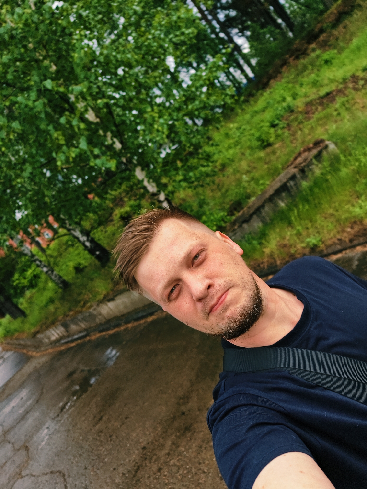 A medium shot captures a man with short, light brown hair and a beard, wearing a dark blue t-shirt and a black strap across his shoulder. He is positioned in the lower right foreground, facing the camera with a slight smirk. The background is tilted, revealing a verdant scene with trees, grass, and what appears to be a concrete structure. A wet, asphalt road runs along the bottom left corner of the frame. The location appears to be in a wooded area within a city, possibly Noviy, Russia, suggested by the architecture and vegetation.