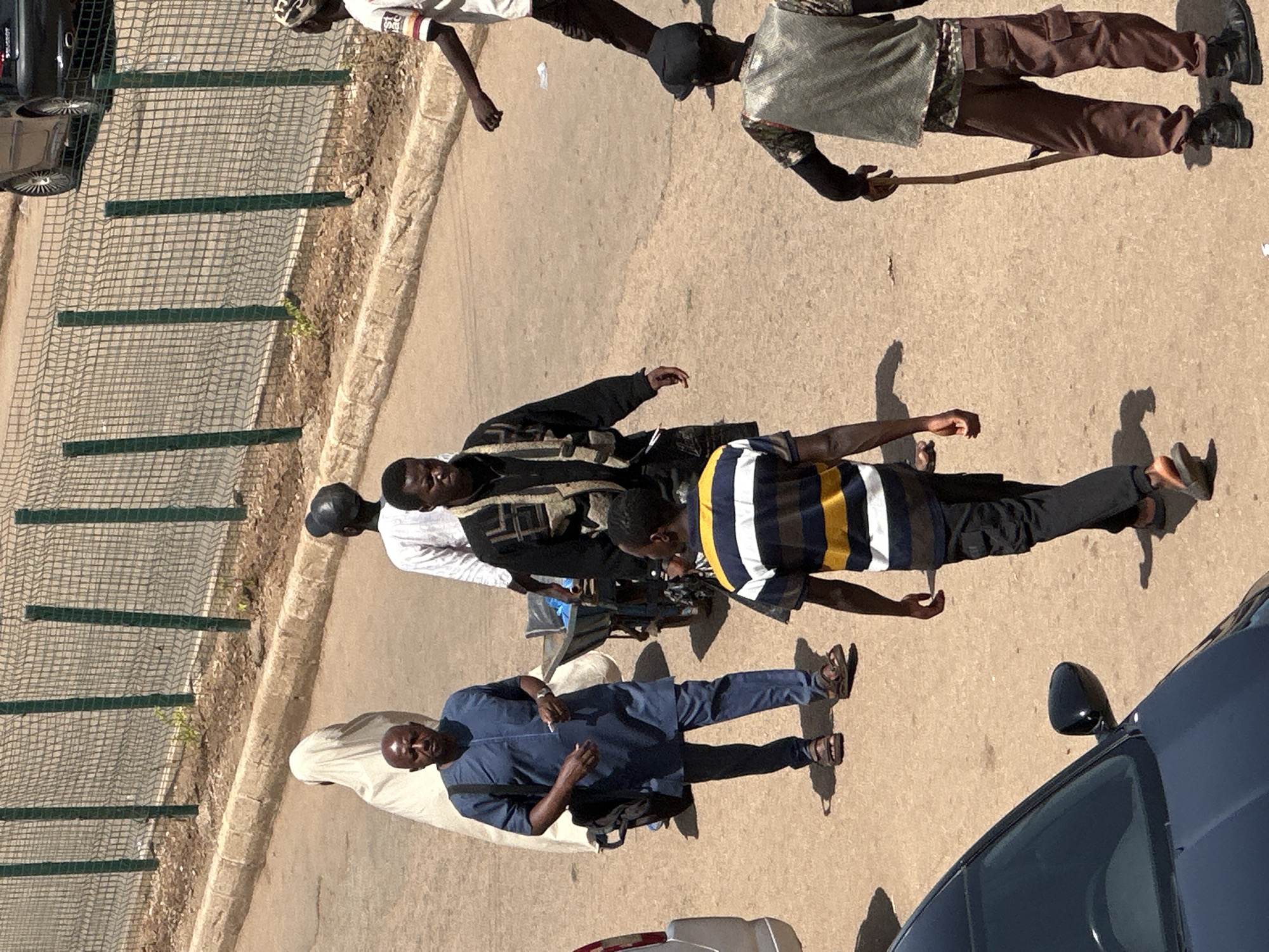 The image depicts an outdoor scene in Bukuru, Nigeria. Several individuals are walking on a paved road. In the foreground, a person wearing a blue tunic and trousers with sandals is walking from left to right. To their left, a person in a white garment is partially visible. Behind the person in blue, another individual is walking with a cart. Further back and to the right, a person in a black jacket over a vest is walking. In the middle ground, a person in a white shirt and dark trousers is walking away from the camera. In the right foreground, a person wearing a patterned vest and brown cargo pants with boots is holding a stick. A dark-colored car is parked on the left side of the frame, and a fence runs along the top of the image, bordering a dirt area with sparse vegetation. The overall lighting suggests it is daytime.