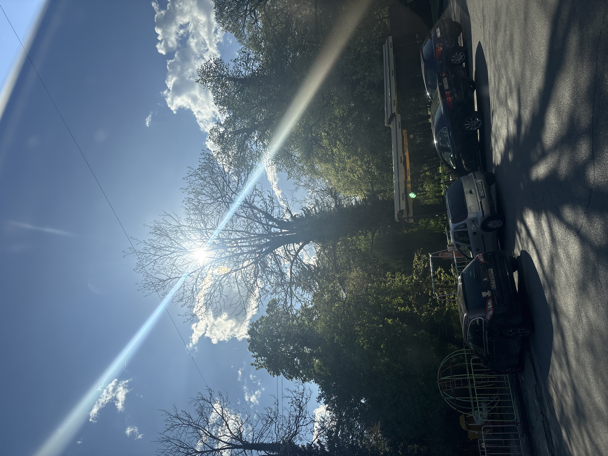 The image shows an upward view of a street in Starozagorski bani, Bulgaria, on a sunny day. Several parked cars are visible along the right side of the street. The sky is blue with scattered clouds, and the sun is partially obscured by a large tree. The light is bright, causing lens flare. Foliage from other trees is visible, along with what appears to be a playground.
