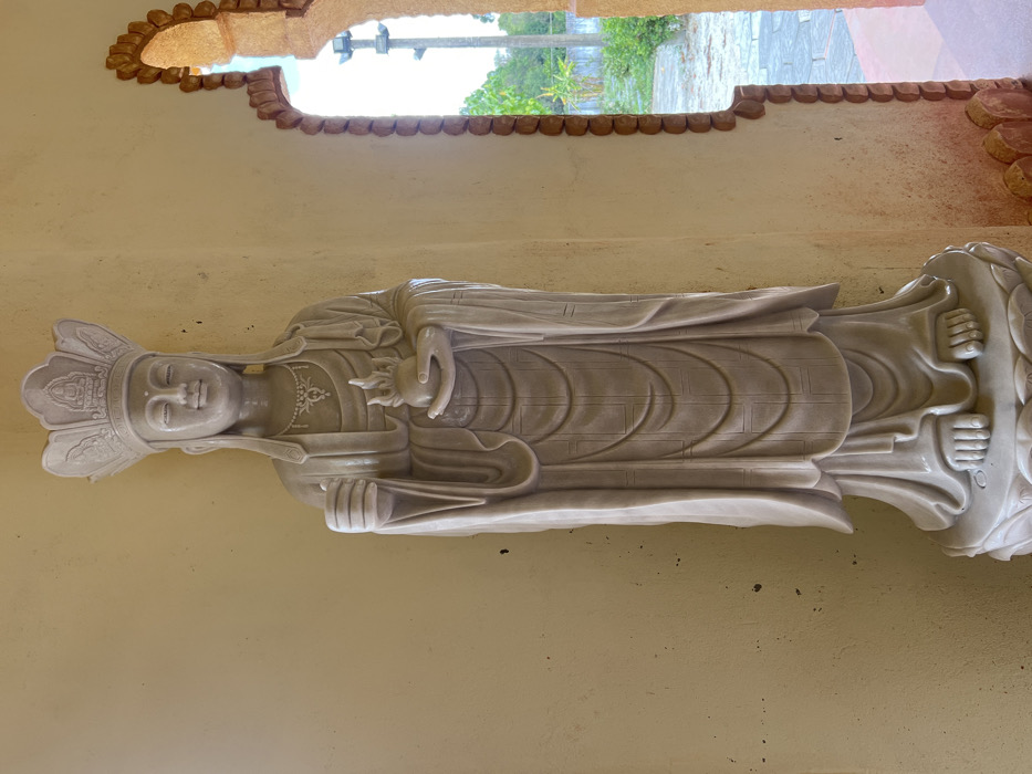 A full-length, pale beige marble statue of a crowned deity stands against a plain, light yellow-beige wall in Đăk Mar, Vietnam. The statue wears a detailed headdress and a necklace, holding an upright cylindrical object in its raised right hand and cradling a flame-shaped object in its left hand. Its flowing robes exhibit subtle linear patterns. The statue rests on a carved lotus-petal base. To the right, an ornate archway with a textured, reddish-brown border frames an outdoor view. This view reveals a paved ground, green foliage, and a utility pole against a bright, clear sky.