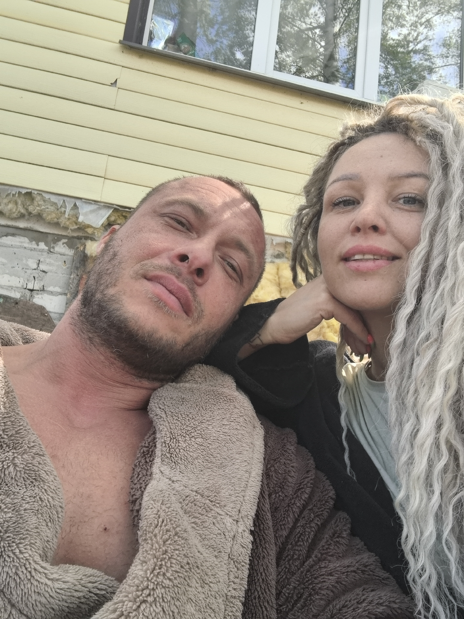 The image captures two individuals, a man and a woman, positioned close together in an outdoor setting. The man, on the left, appears to be wearing a brown, textured bathrobe. His chest is partially visible, and he has short hair and a beard. The woman, to the right, has light-colored dreadlocks and is wearing a dark robe over a light-colored shirt. Her hand is placed under her chin.
Behind the subjects is a wall with light yellow siding and a window. Insulation material is visible on the wall. Based on the presence of Russian characters, the location is in Russia. The city is Noviy. The image appears to be a selfie or taken at close range, with the focus on the subjects' faces.