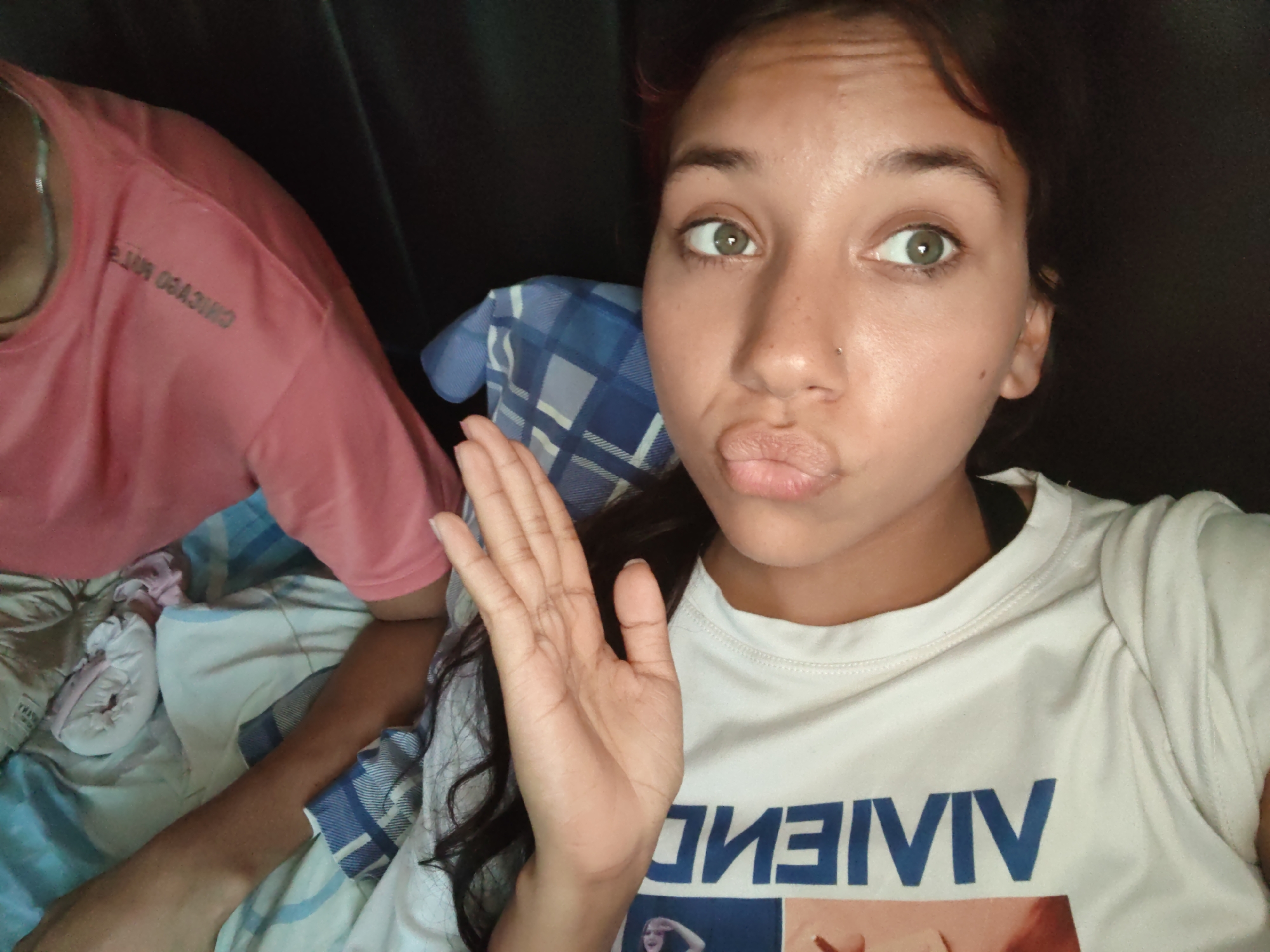In Piloncito, Venezuela, a young woman with dark hair and green eyes is shown in a close-up, selfie-style photograph. She is wearing a white t-shirt with large blue lettering that reads "VIVIEN." Her mouth is puckered as if she is about to kiss, and her left hand is raised in a welcoming gesture. To her left, another person is visible wearing a pink t-shirt. The background consists of a dark surface, possibly furniture, and bedding with a blue and white plaid pattern. The lighting is soft, highlighting her facial features.
