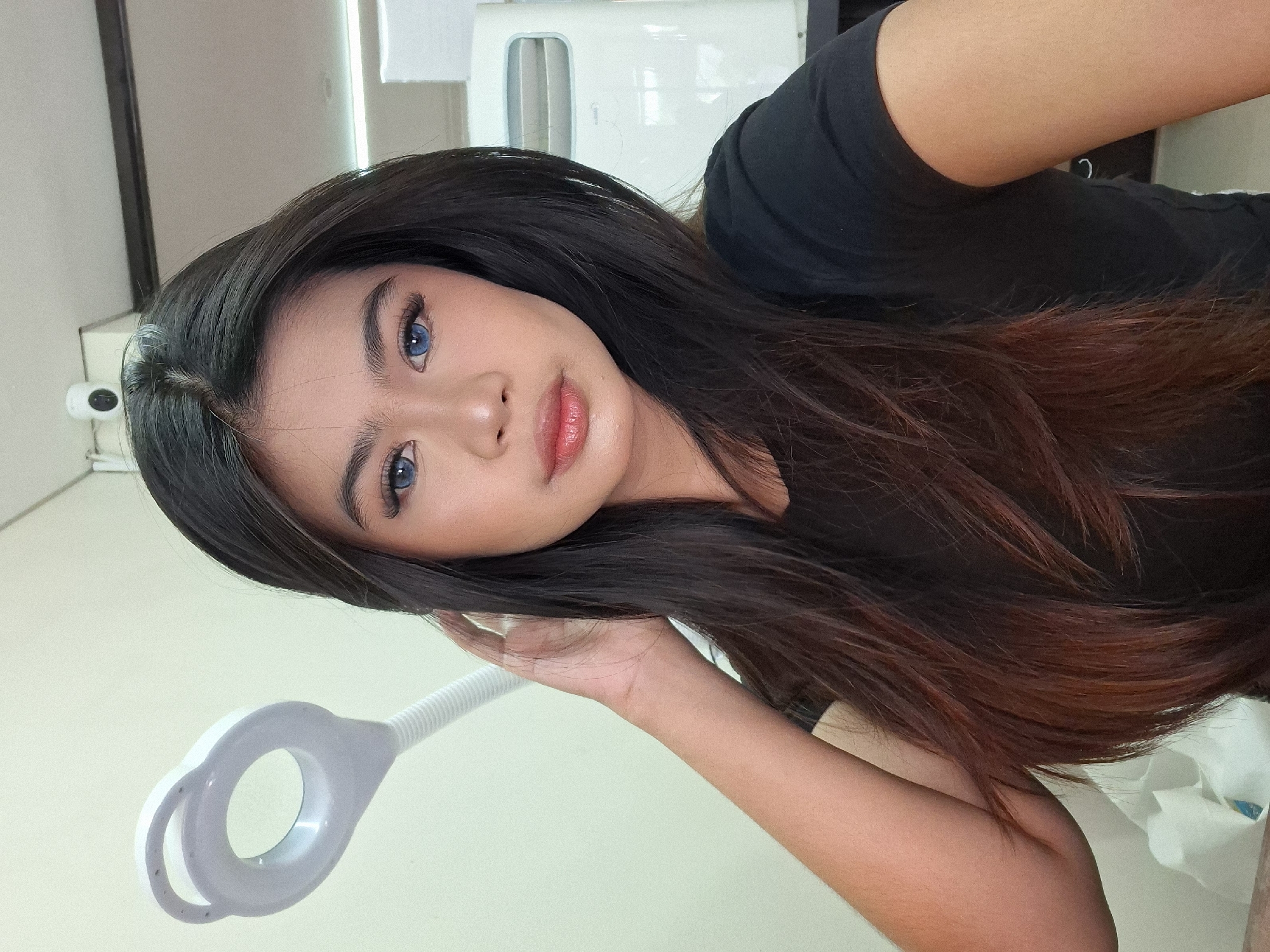 A young woman with long, dark brown hair is captured in a selfie. She has blue contact lenses and is wearing makeup. Her right hand is raised to her head, and she is looking directly at the camera. She is wearing a black t-shirt. In the background, a white magnifying lamp with a ring light is visible to the left. A white electrical appliance with a gridded front panel is to the right, and a small security camera is mounted on the ceiling. The walls are a light, neutral color. The scene appears to be indoors, likely in a beauty salon or similar setting in Kuta, Indonesia.