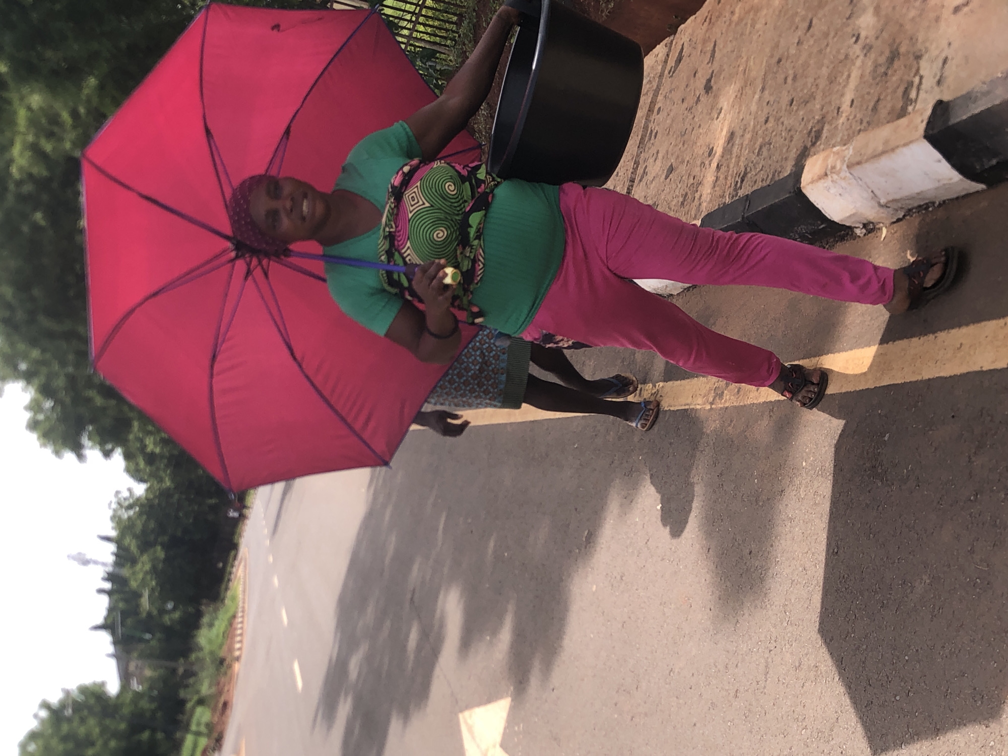 A woman is standing on a paved road in Nsugbe, Nigeria. She is holding a red umbrella over her head. She is wearing a green shirt, pink pants, and sandals. She has a patterned wrap around her torso, possibly holding a baby or other item. In her left hand, she is holding a large black bucket. The woman is smiling.
Another person, possibly a child, is standing behind her to the left. This person is wearing a patterned skirt and sandals.
The background includes trees and a building. The road is marked with a solid yellow line. Along the edge of the road is a curb painted white and black. The image appears to be taken during the daytime.
