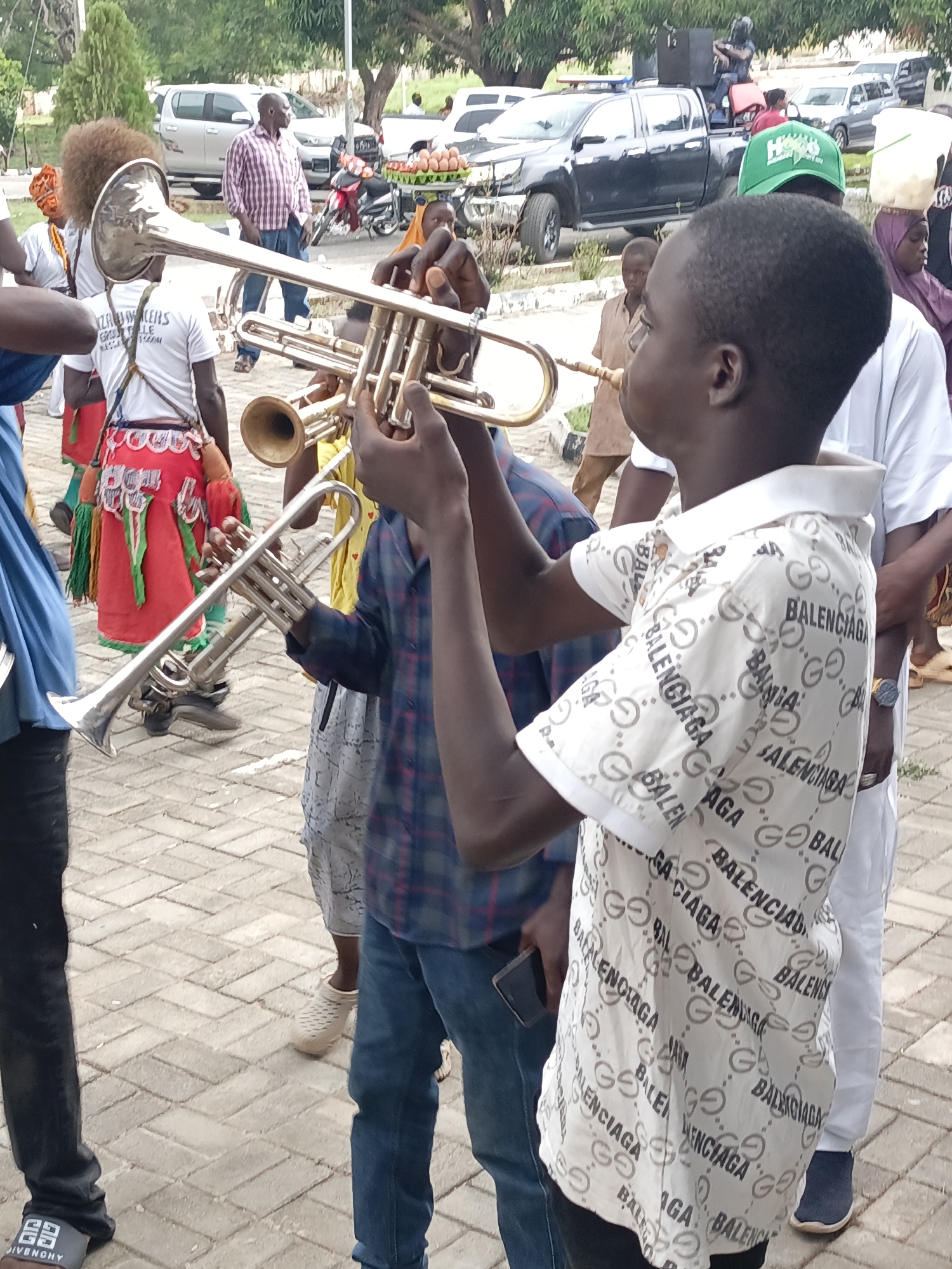 The image shows a group of people in what appears to be an outdoor gathering in Akwanga, Nigeria. Several individuals are holding and seemingly playing trumpets. A young person in the foreground is wearing a white short-sleeved shirt with "Balenciaga" printed repeatedly. Other individuals are wearing traditional attire, including a red skirt. In the background, there are parked cars and a pickup truck with a person sitting on top of it. The ground is paved with interlocking blocks.