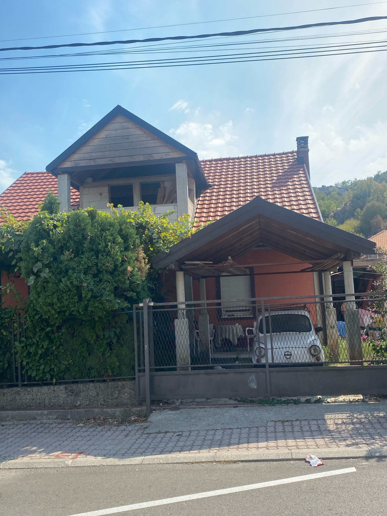 The image depicts a residential property in Cetinje, Montenegro. The scene shows a two-story house with a red tile roof. The upper part of the house appears to be under construction or unfinished, with exposed wooden beams and concrete structures. In front of the house, a carport covers a light-colored, vintage compact car, possibly a Fiat 500. The property is enclosed by a metal fence. A paved sidewalk runs along the front of the property, adjacent to a road with a white line marking. Lush green foliage and bushes are visible around the house. The sky is blue with scattered clouds, and a chain of power lines runs across the top of the frame. The overall environment suggests a sunny day in a residential area.