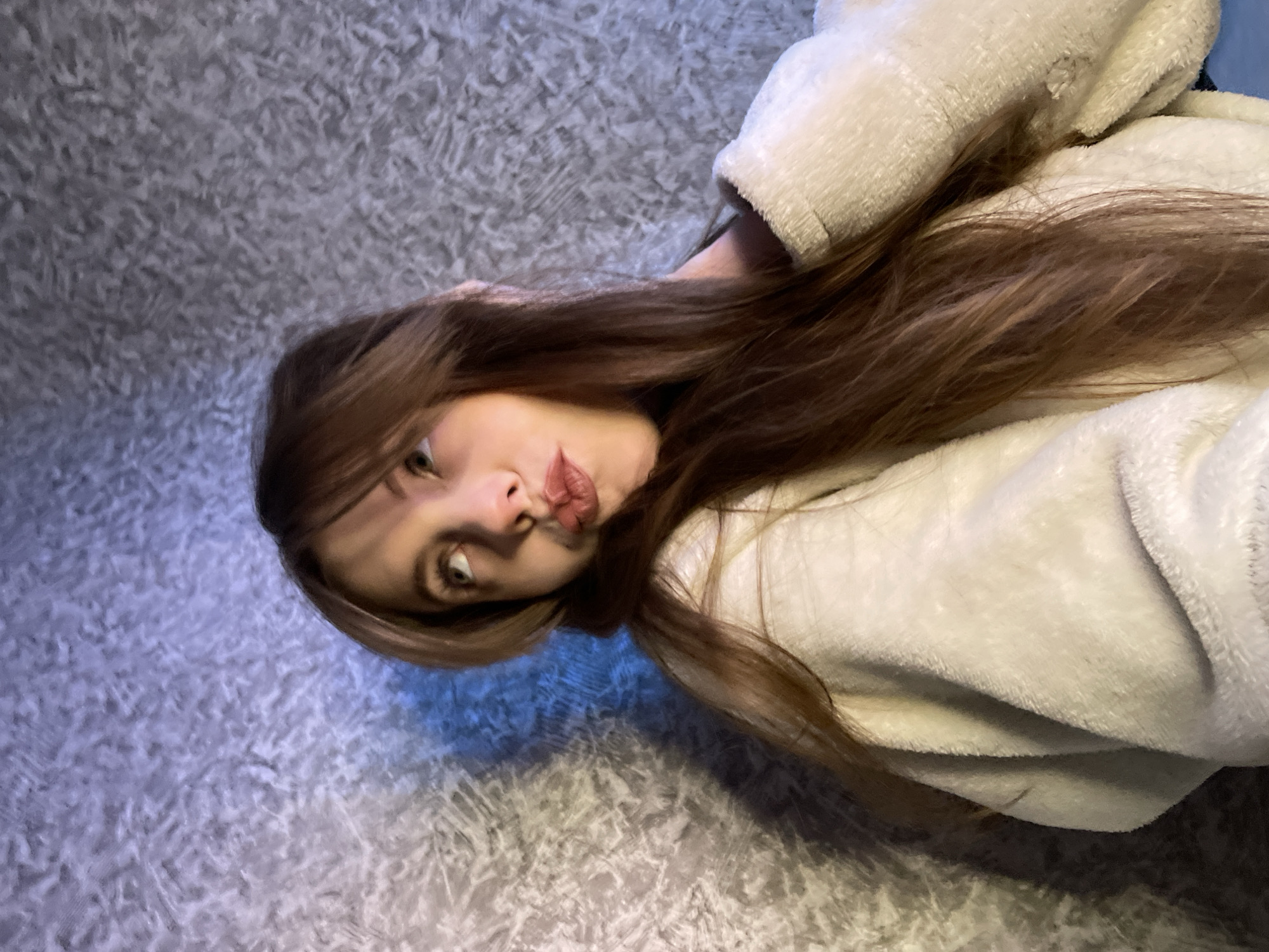 A young woman with long brown hair and fair skin lies on a gray, textured surface, possibly a rug or carpet. She wears a light-colored, fluffy hooded sweatshirt or robe. She is looking directly at the camera with a slight pout or kissing expression. The environment appears to be indoors, illuminated with a mix of natural and artificial lighting. There is no indication of the location being in the city of Фоки, Russia, or any other location-specific identifiers visible in the image.