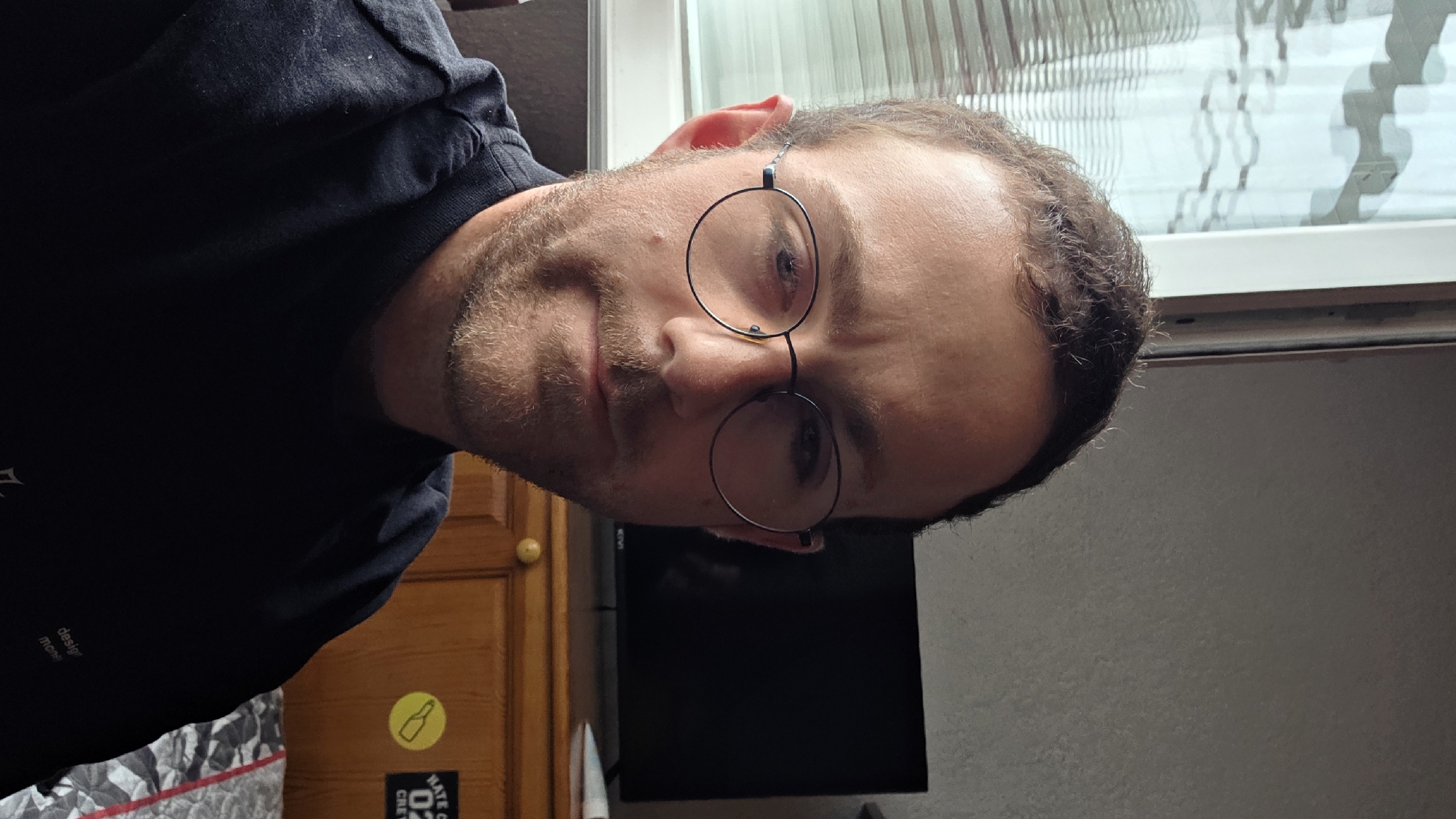 The image is a portrait of a man, presented at a 90-degree counter-clockwise rotation. The man, positioned centrally, has short brown hair and a light brown beard. He wears round, thin-rimmed dark glasses and a dark, possibly navy or black, short-sleeved t-shirt. His expression is neutral as he looks directly towards the viewer.
The background suggests an indoor setting. To the man's left (when the image is correctly oriented), a light brown wooden cabinet or door with a round, gold-colored knob is visible. Below this, a grey and white patterned blanket or textile is partially seen. In the center-bottom, a dark, rectangular screen or monitor is present. To the man's right, a window with a translucent, textured pattern or blinds is partially visible, allowing in soft, diffused light, suggesting daylight.
Visible text includes faint white lettering on the lower left side of his t-shirt, which reads "design" and "more" or "mons." Affixed to the wooden surface near the bottom is a circular yellow sticker featuring a black outline of a bottle. Below it, a black rectangular sticker displays white text: "NATE CA", "02", and "CREW". The overall atmosphere is casual. The location context provided is Bania, Bulgaria.